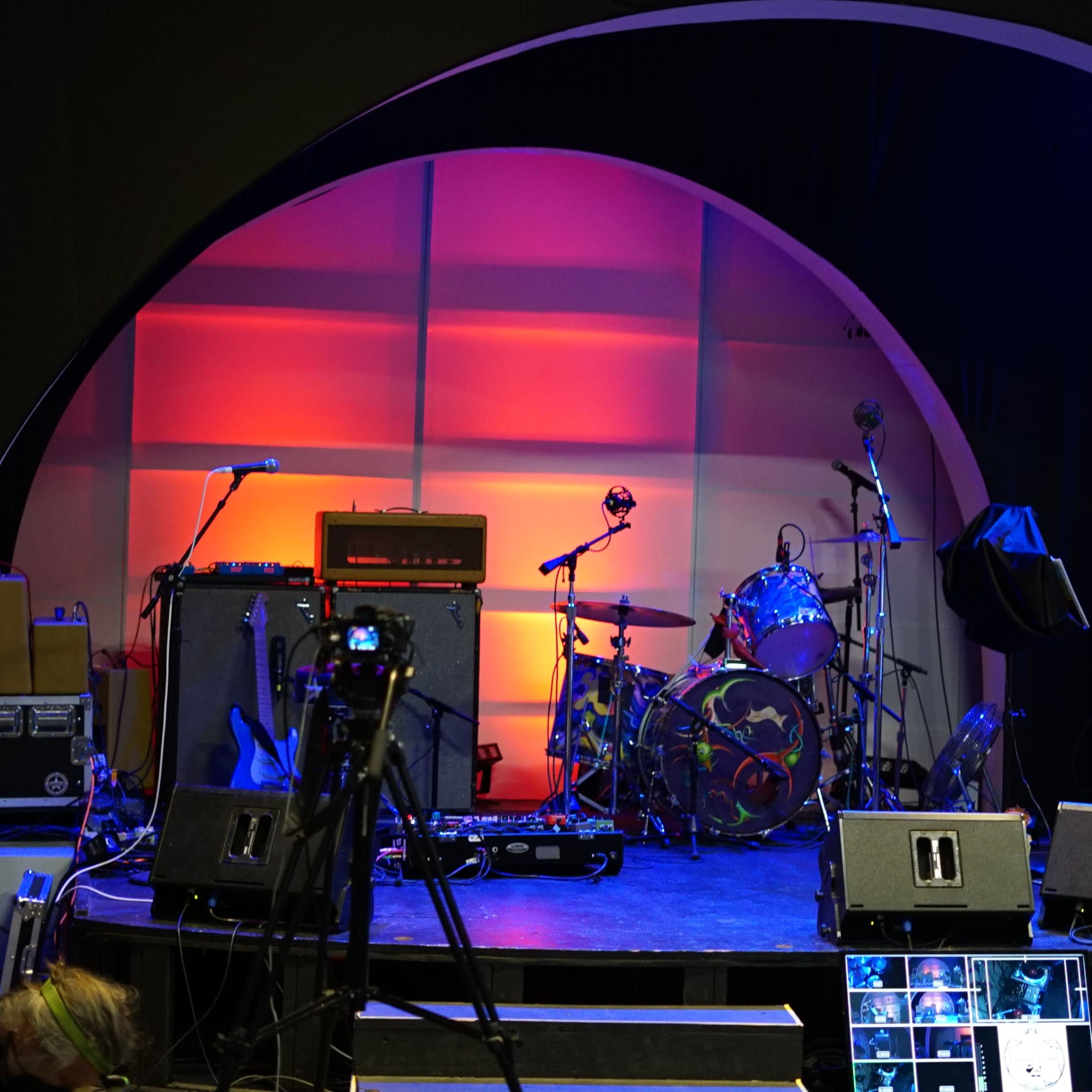 A stage setup with musical instruments, amplifiers, and cameras arranged in a semi-circular backdrop.
