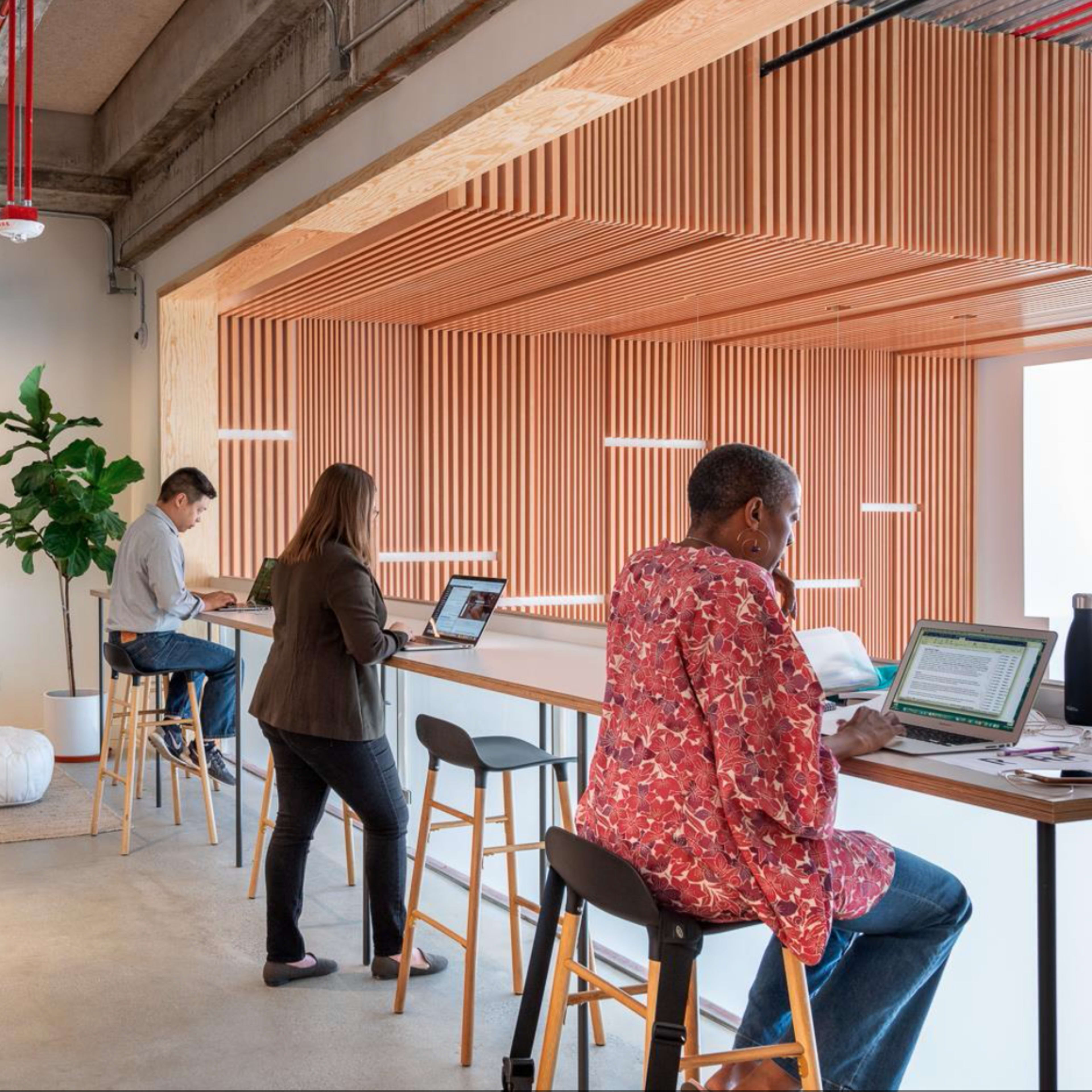 A modern office space with people working on laptops, seated at a communal table and on stools, surrounded by plants and wooden decor.