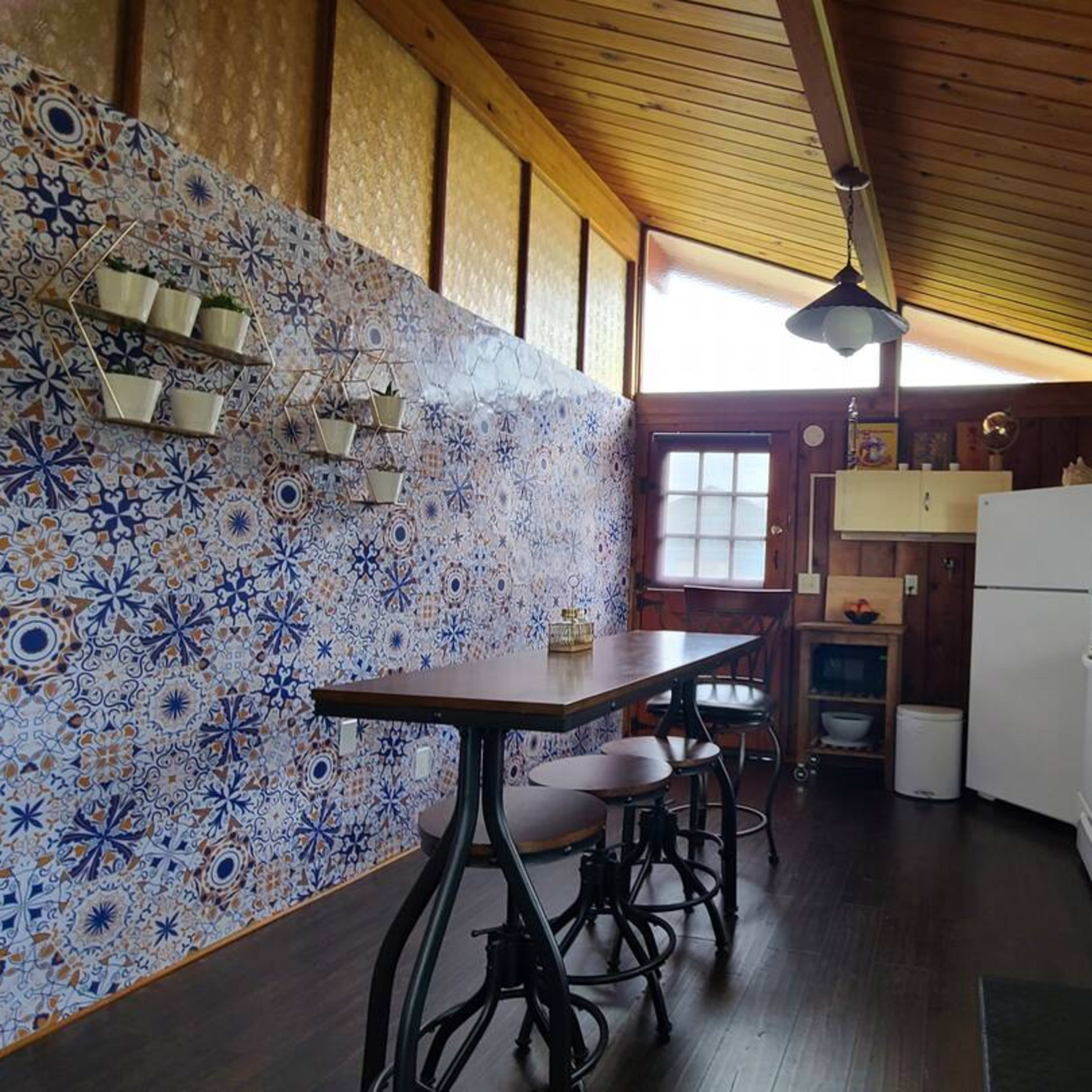 A kitchen with a tile accent wall, a wooden ceiling, a white fridge, a sink with a window, and a dining table with three stools.