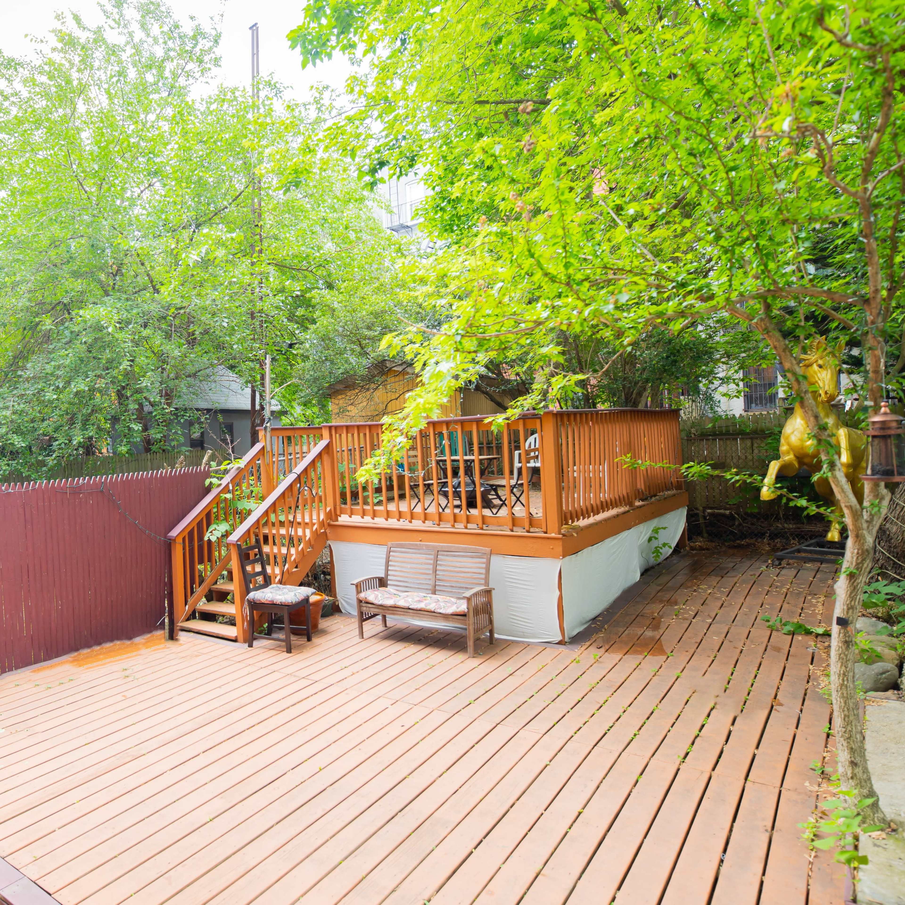 A wooden deck with a staircase leading to a raised area surrounded by greenery and a red fence.