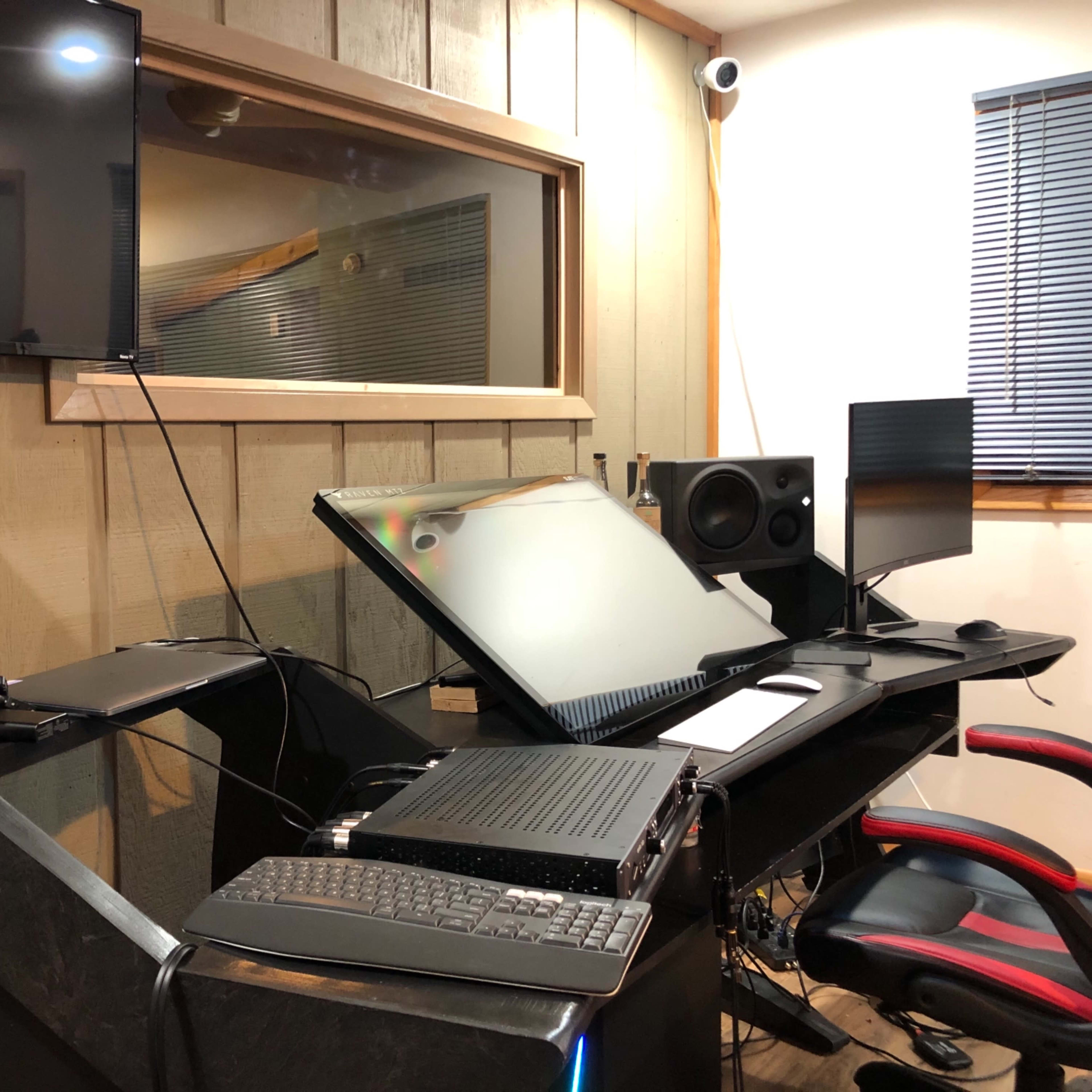 A music studio setup with a large mixing console, multiple monitors, speakers, and an ergonomic chair against a wooden-paneled wall.