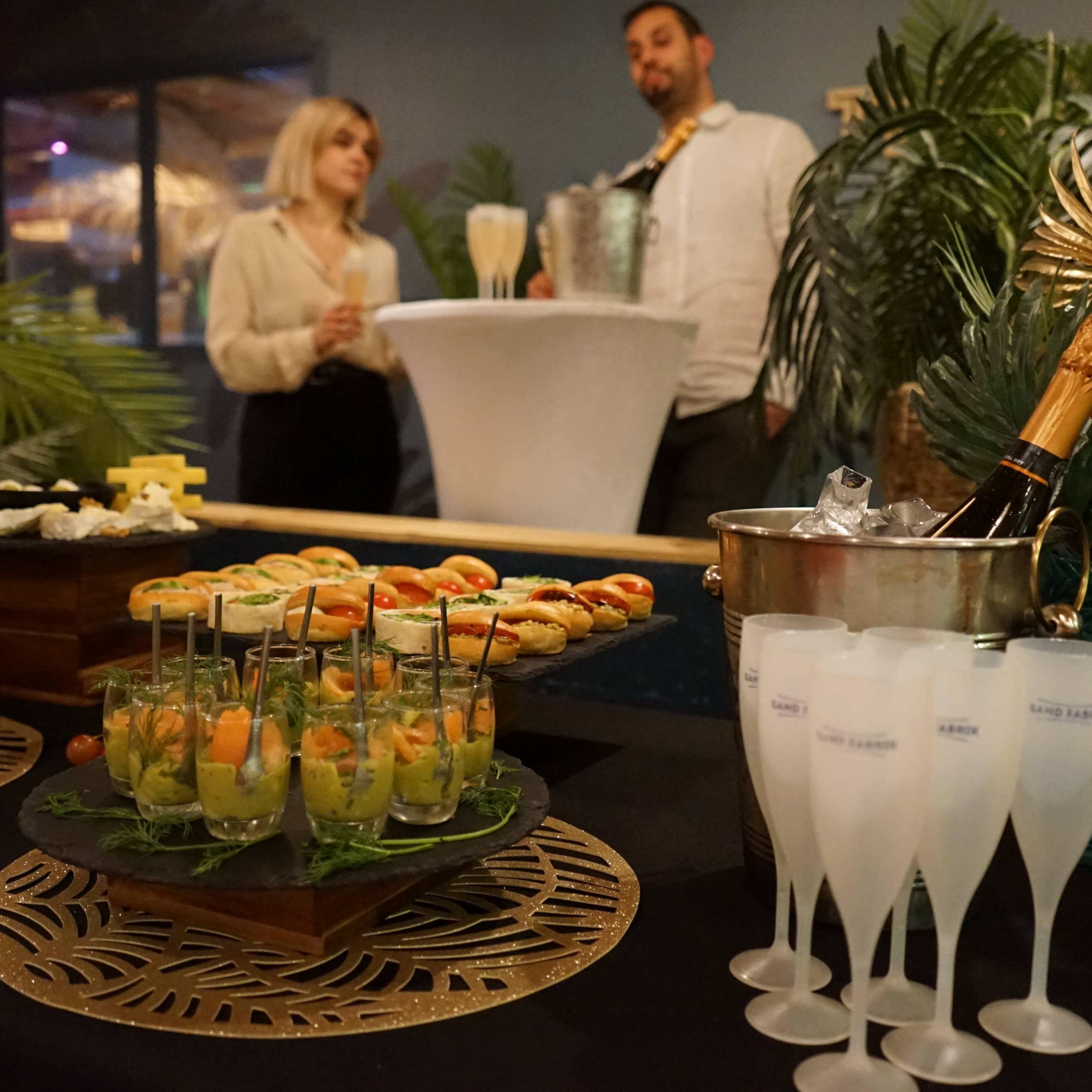 A table is set with various appetizers and champagne flutes, while two people stand nearby, one holding a bottle of champagne.