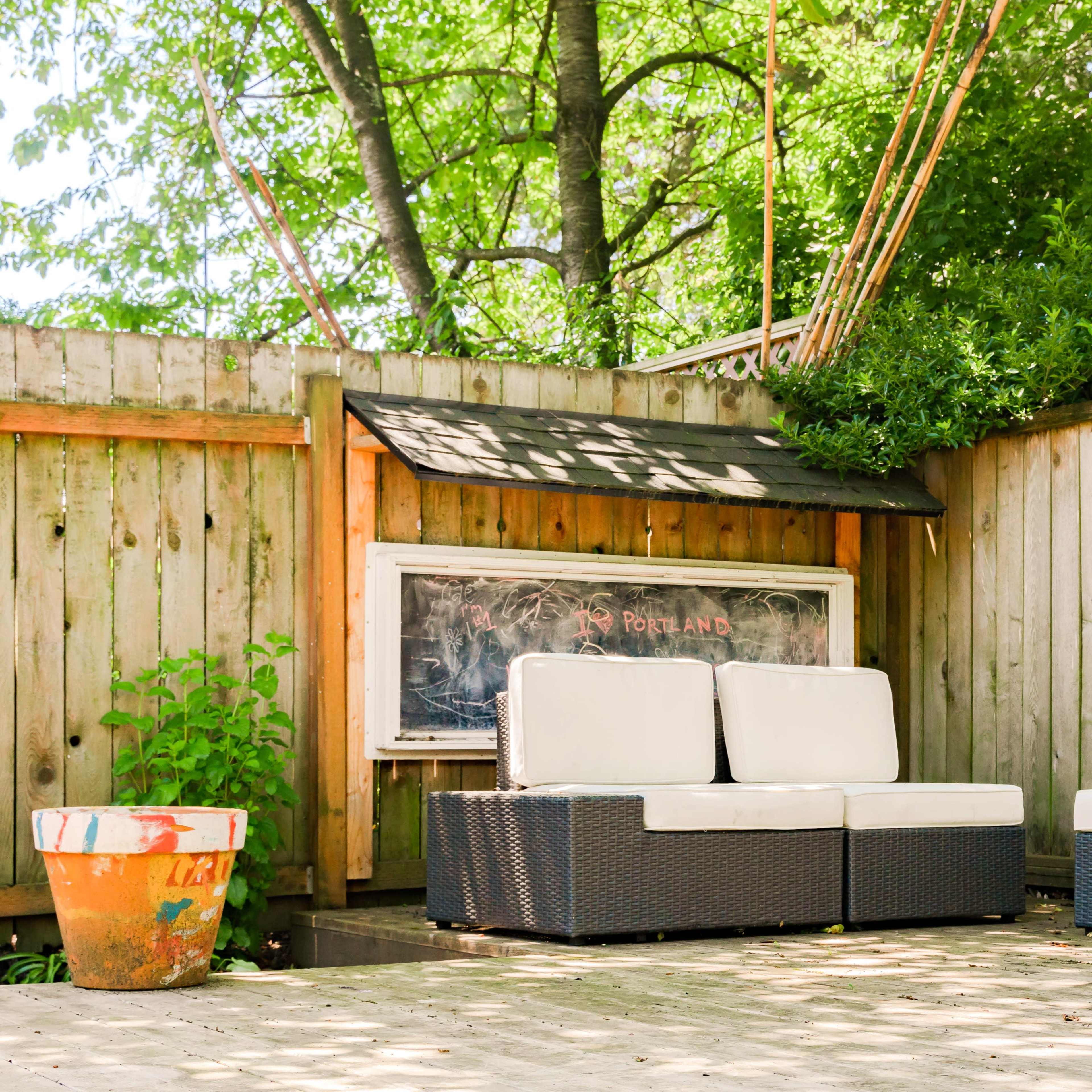 A seating area with white cushions on dark wicker furniture is set against a wooden fence with a chalkboard and a potted plant nearby.