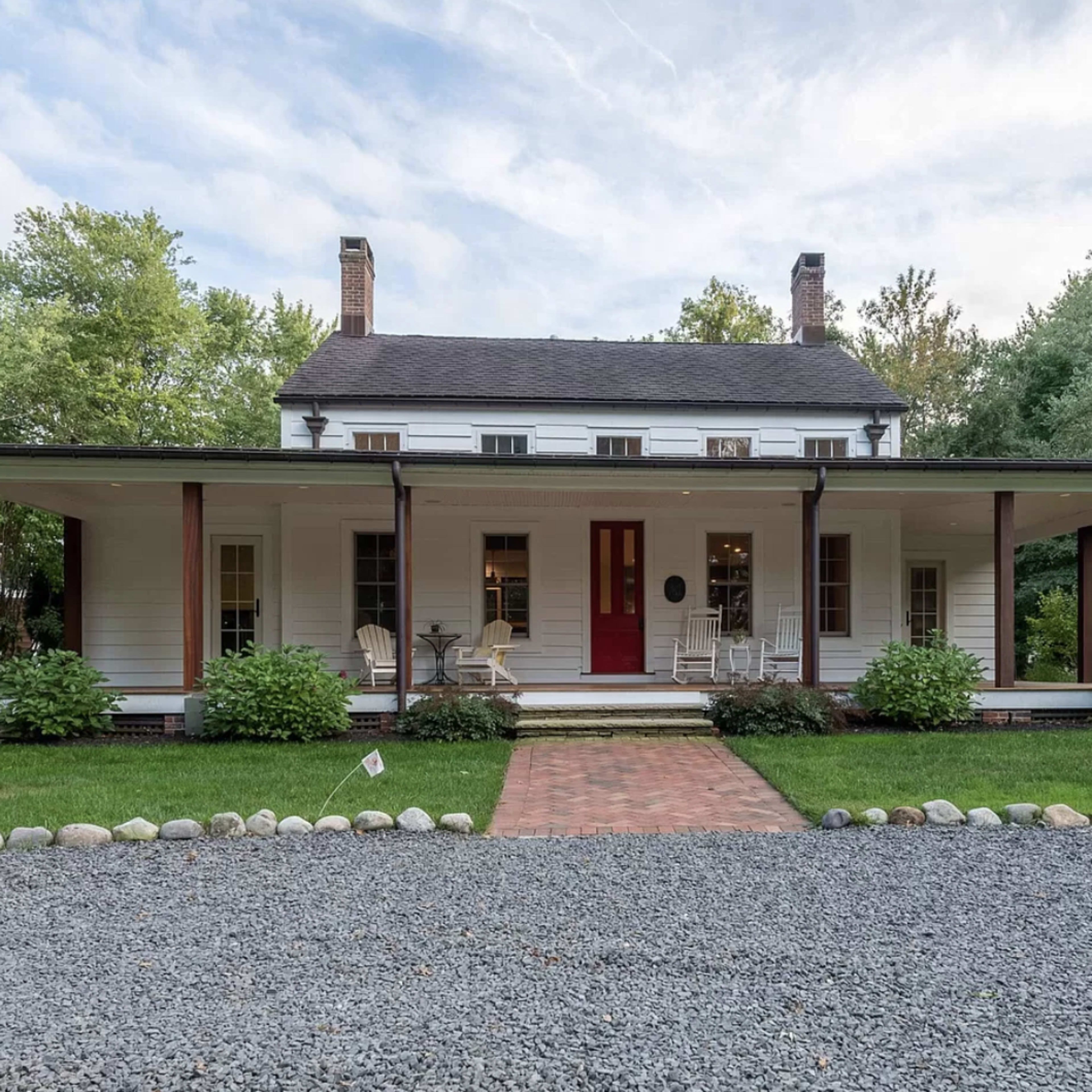 A two-story, white wooden house with a red door, surrounded by a green lawn, gravel driveway, and landscaped bushes.