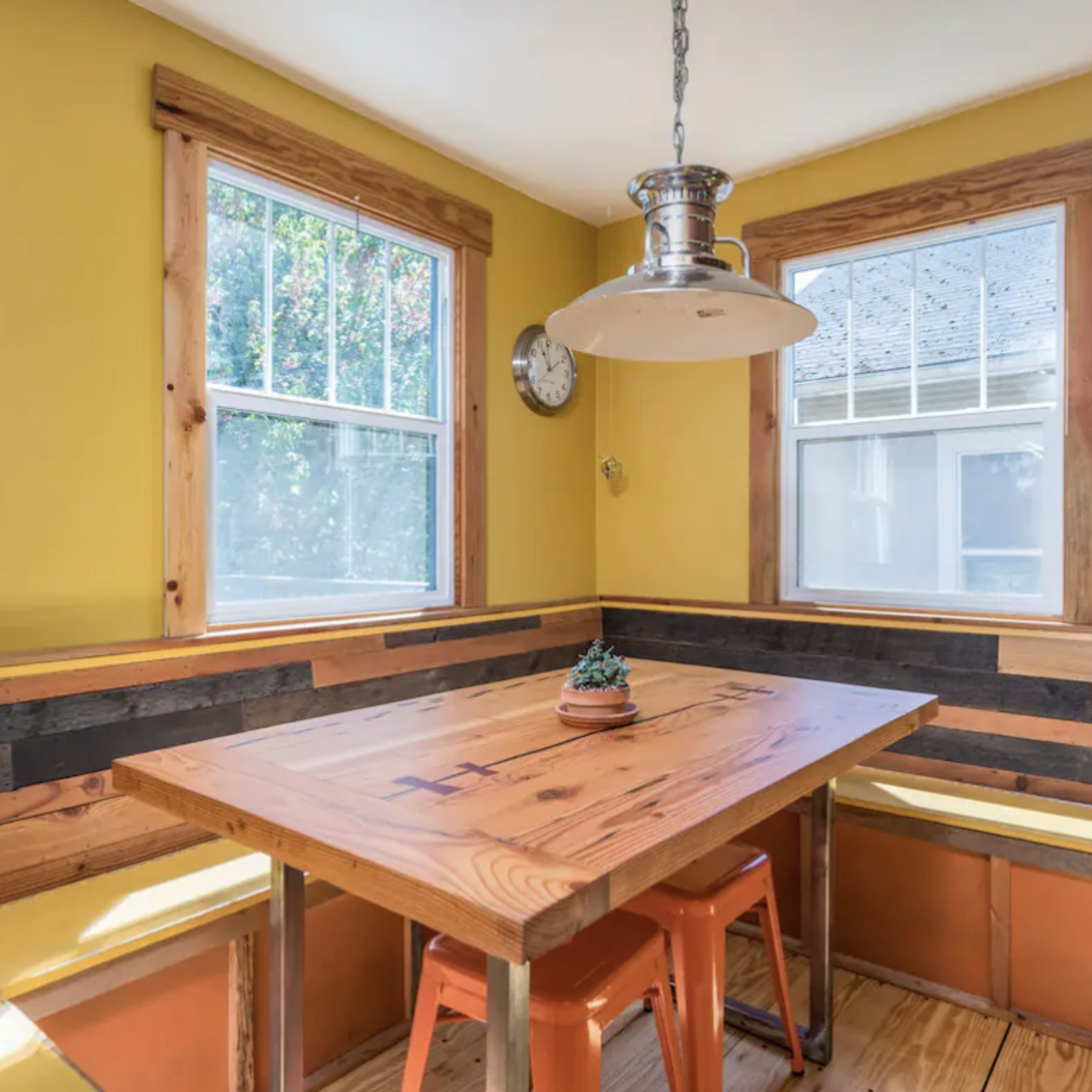 A dining area features a wooden table surrounded by benches and chairs, with yellow walls and large windows allowing natural light to enter.