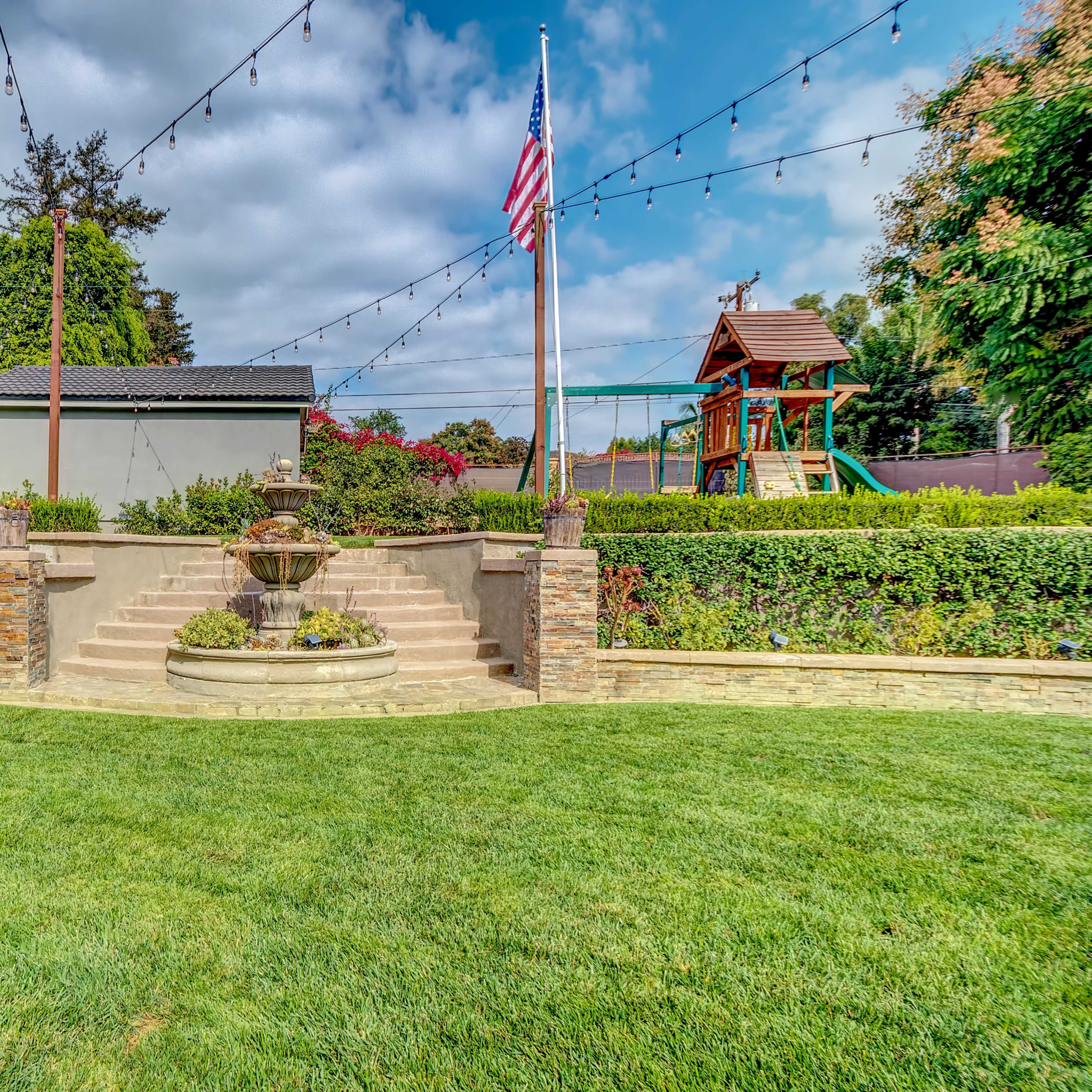 A landscaped backyard featuring a central stone fountain, a grassy area, and a children's play structure in the background, with string lights and an American flag.
