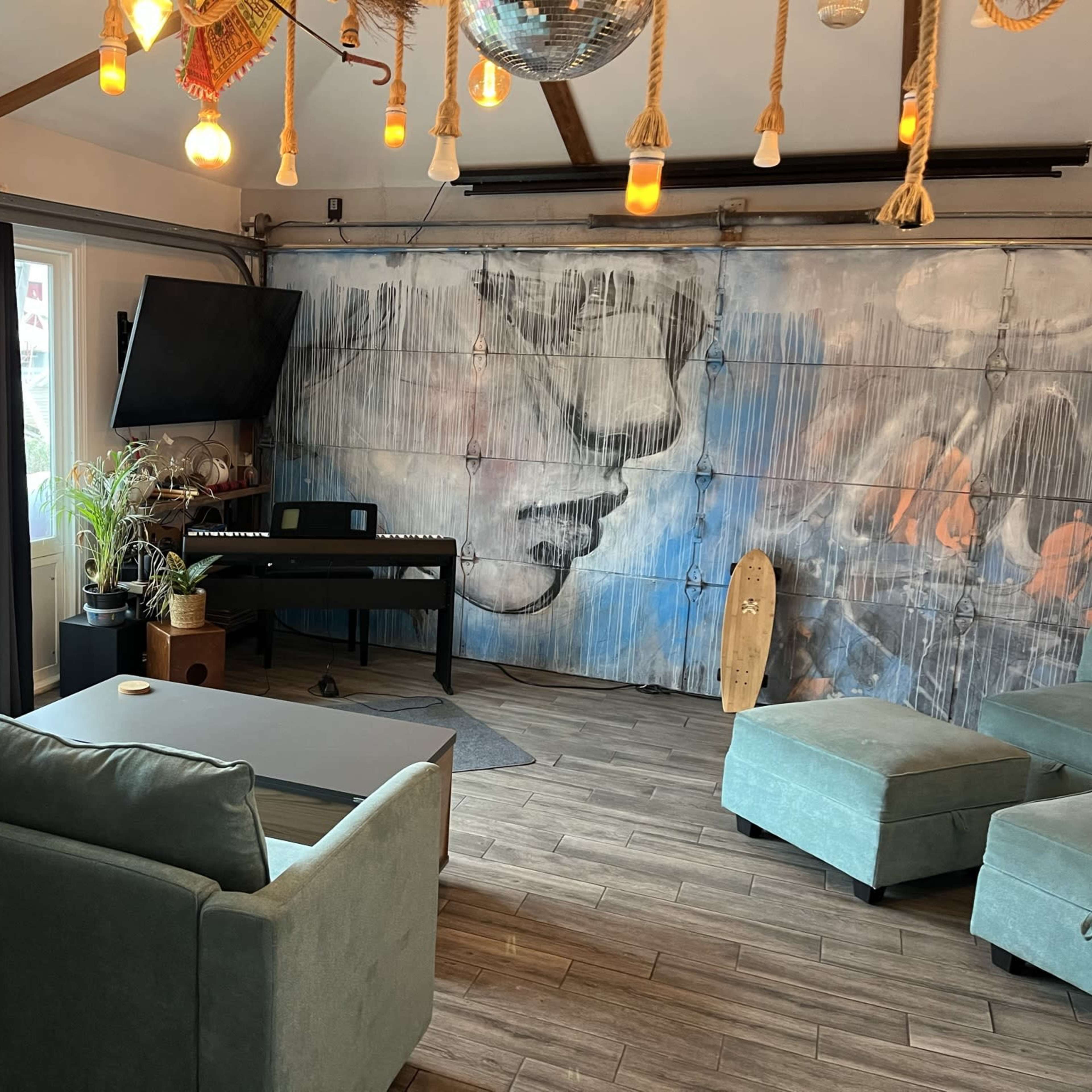 The room features a large mural of a face on one wall, a digital piano, a couch, and pendant lights hanging from the ceiling.
