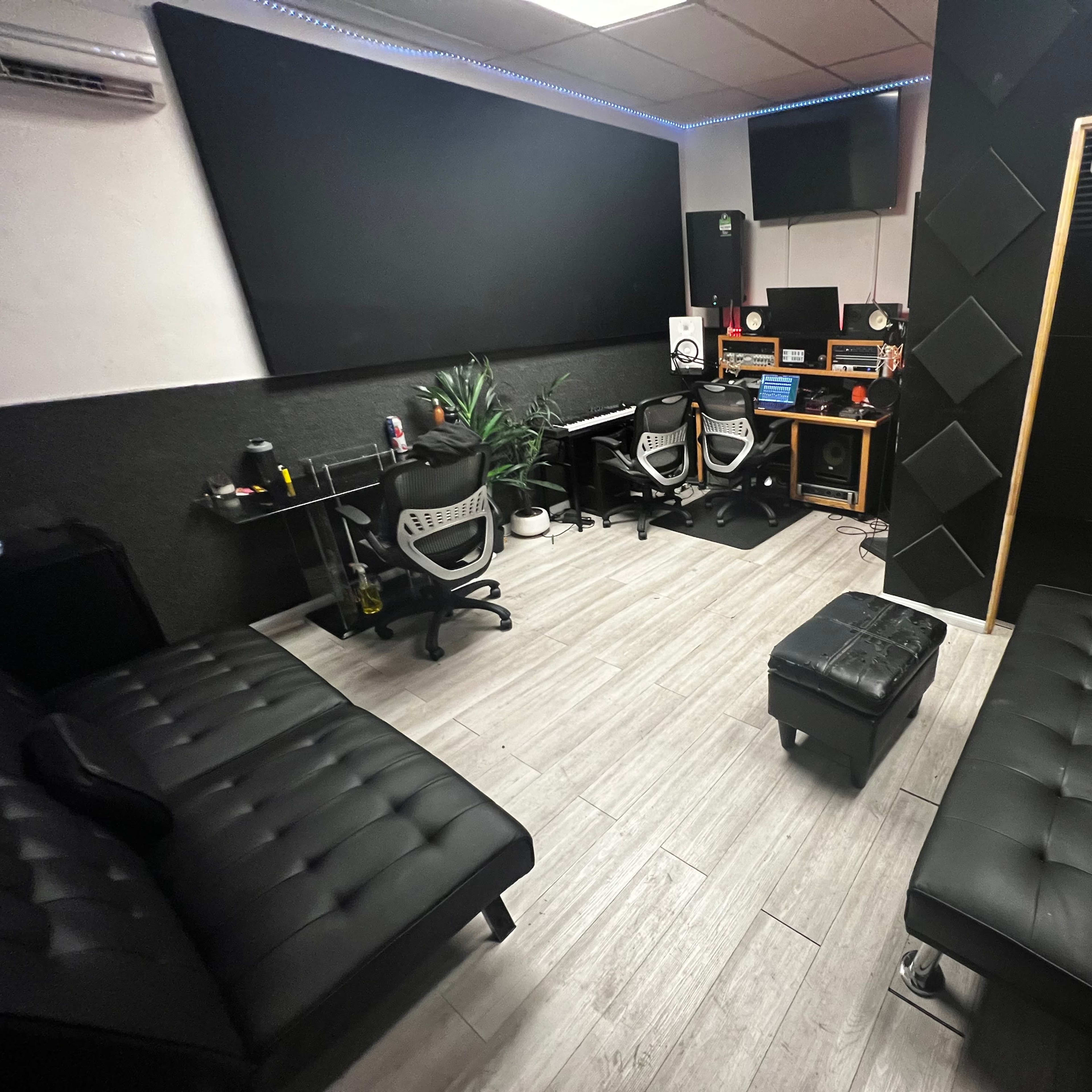 A modern recording studio featuring black sofas, a work desk with audio equipment, and a soundproof door.
