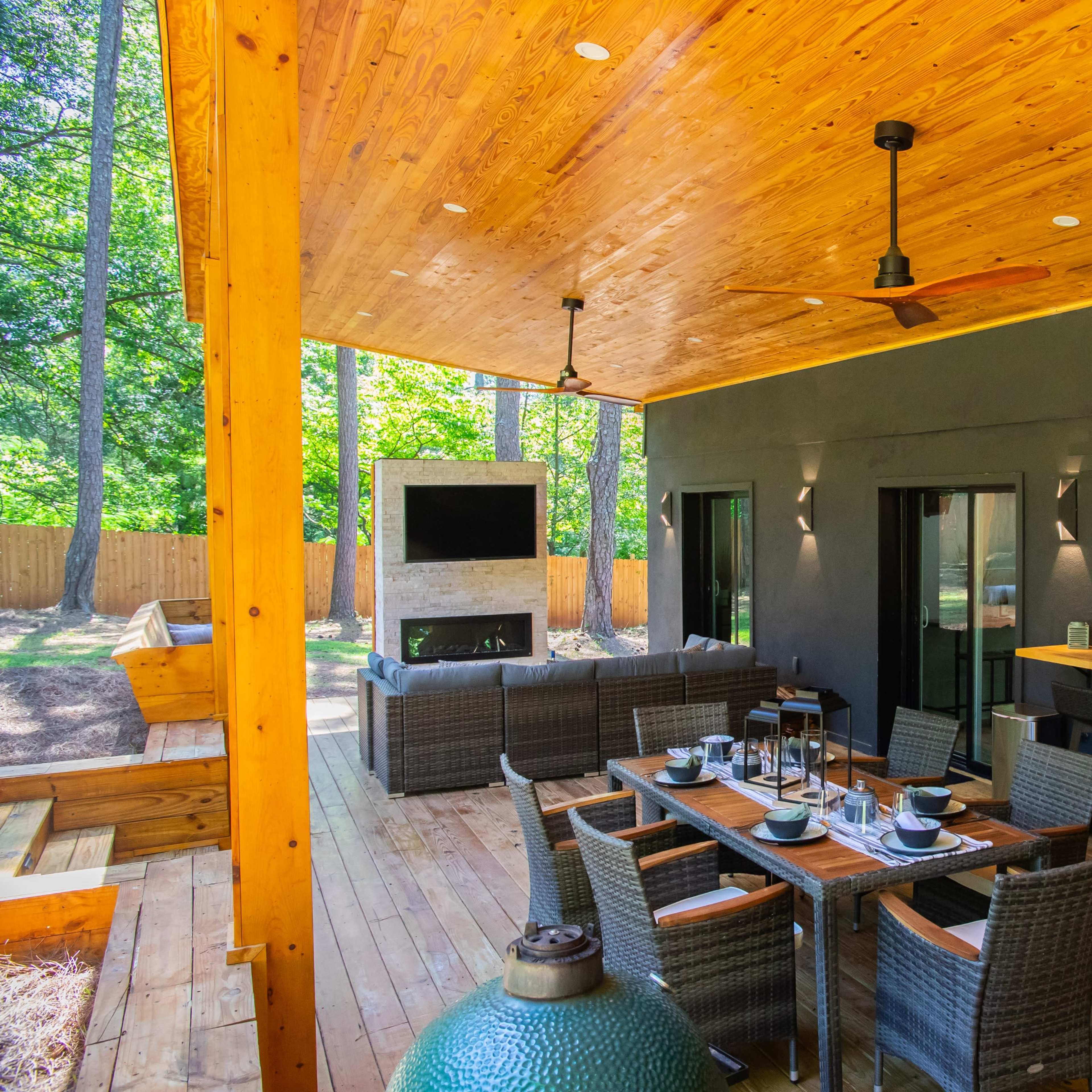 A spacious covered patio with a dining area, seating arrangement, and a large outdoor television, surrounded by trees.