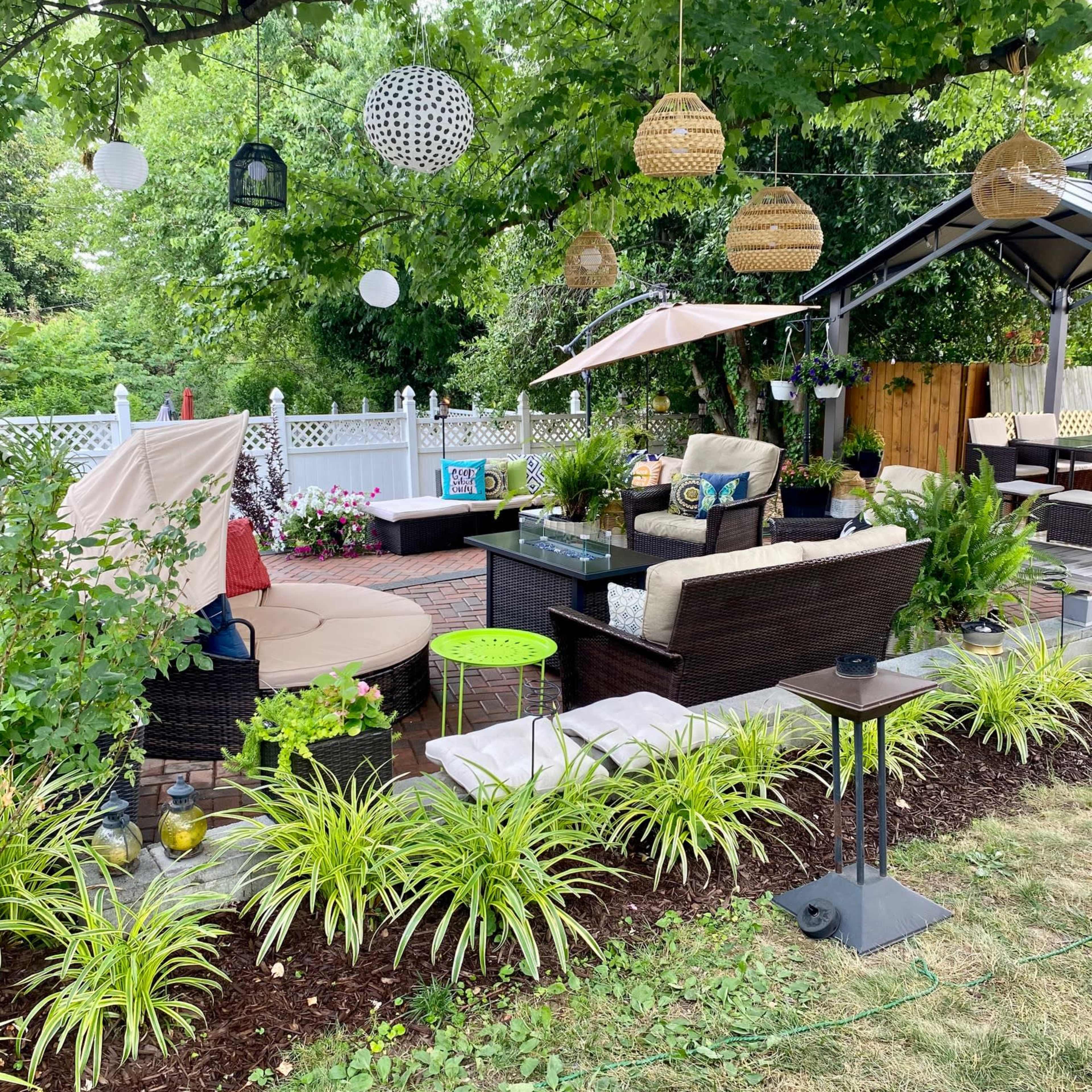 A landscaped outdoor seating area with patio furniture, decorative hanging lights, and potted plants under a large tree.