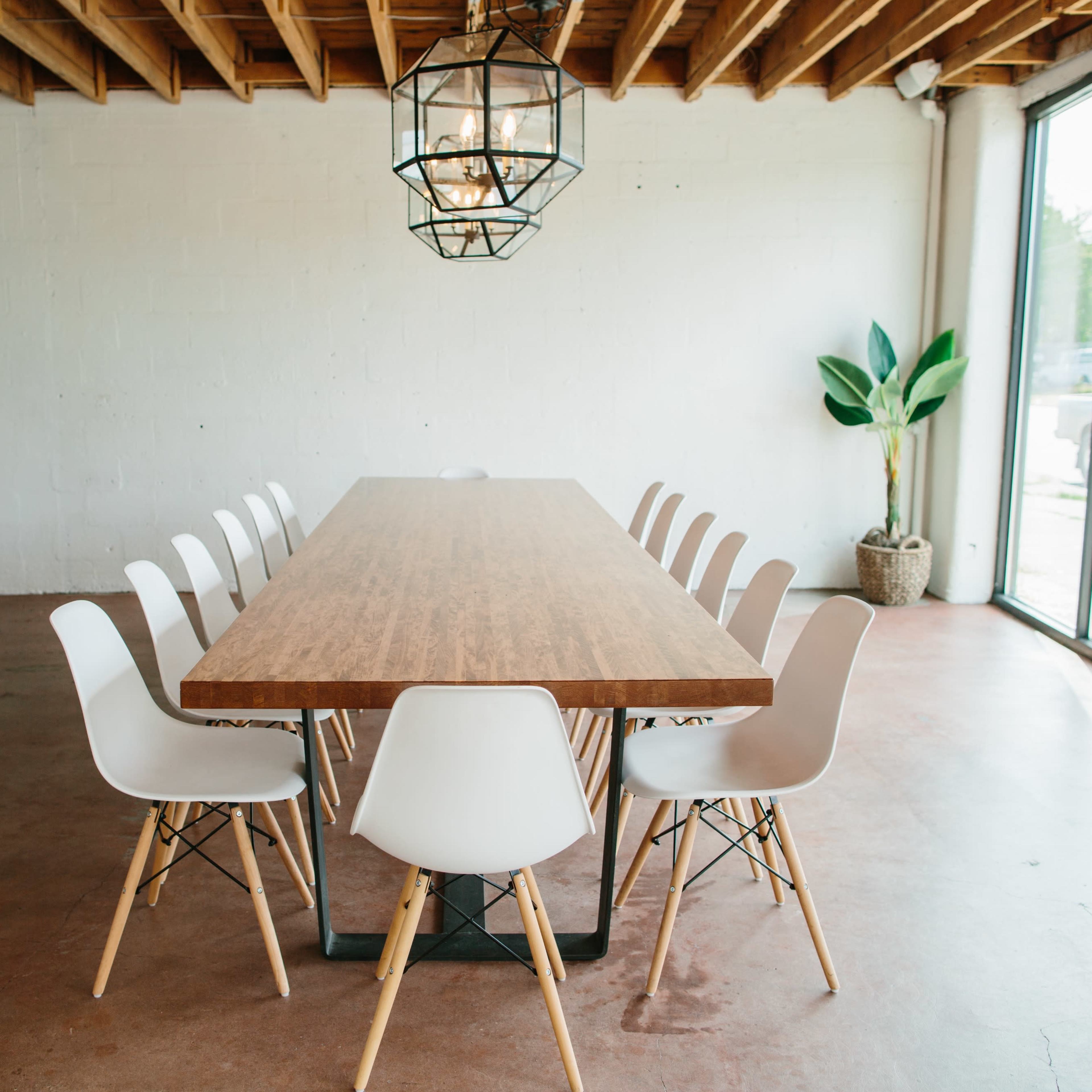 A long wooden table with white chairs is set in a spacious room with large windows and a pendant light fixture.