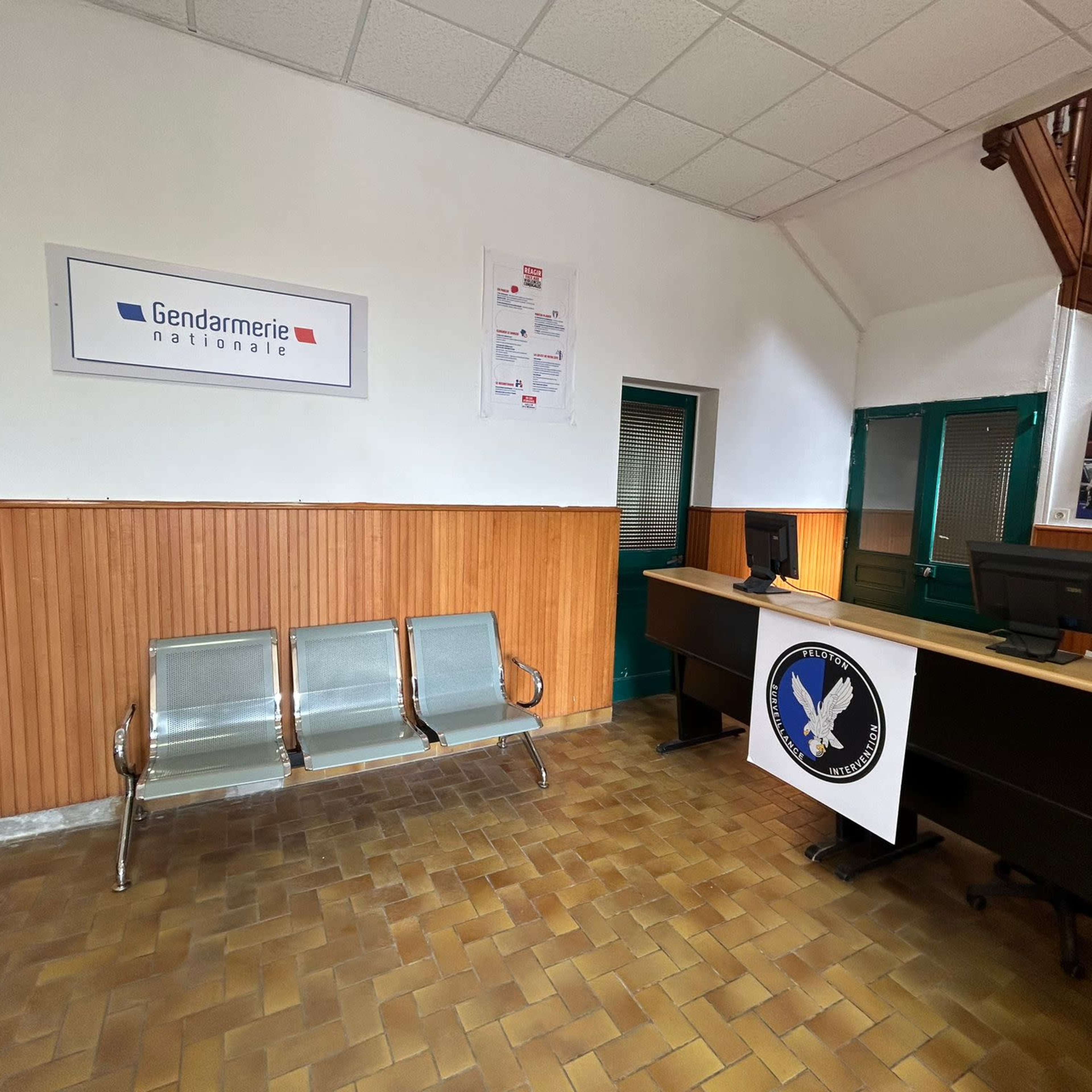 The interior of a gendarmerie office, featuring a reception desk with computers and metal benches against a wooden wall.
