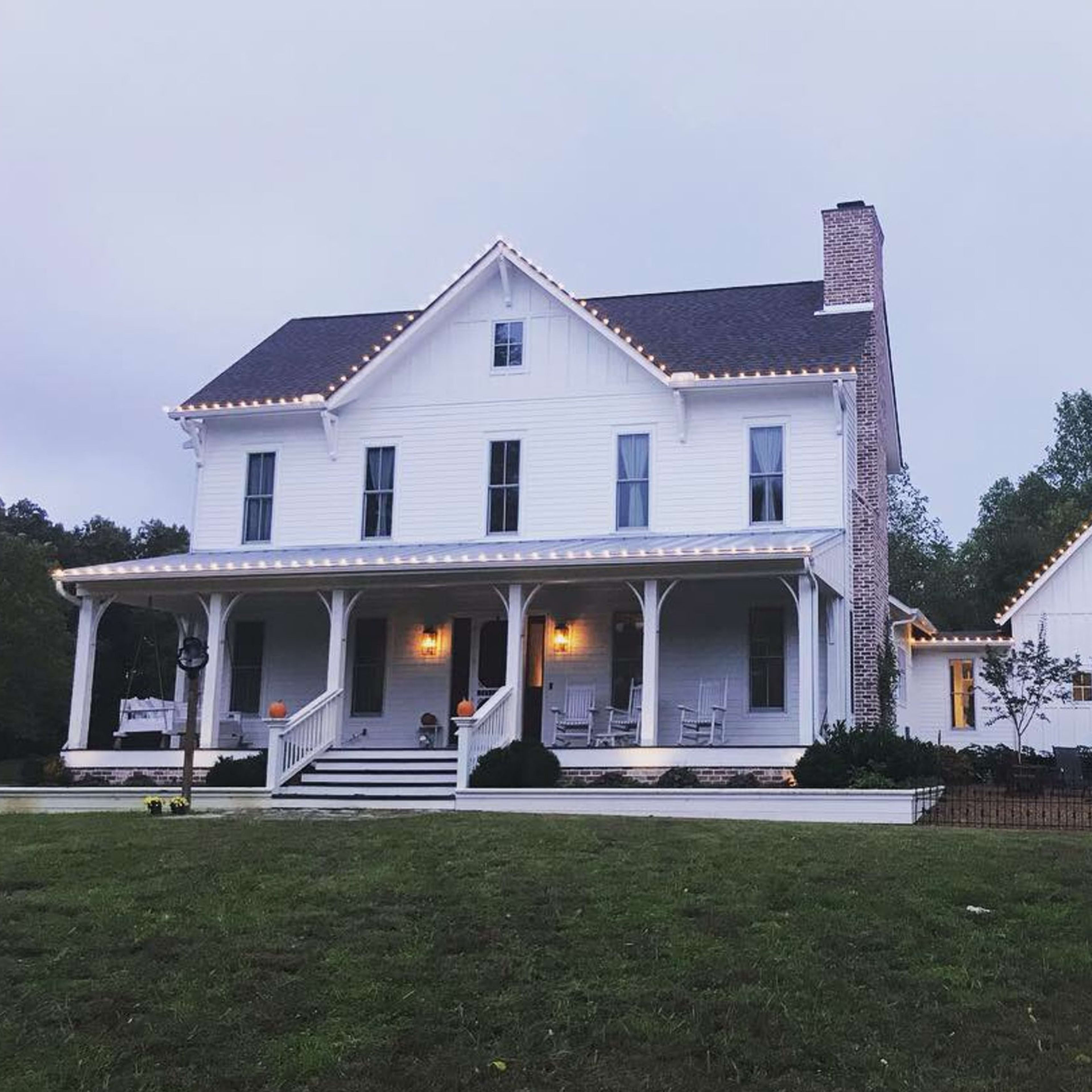 A two-story white house with a gabled roof features string lights along the eaves and a porch surrounded by a landscaped yard.