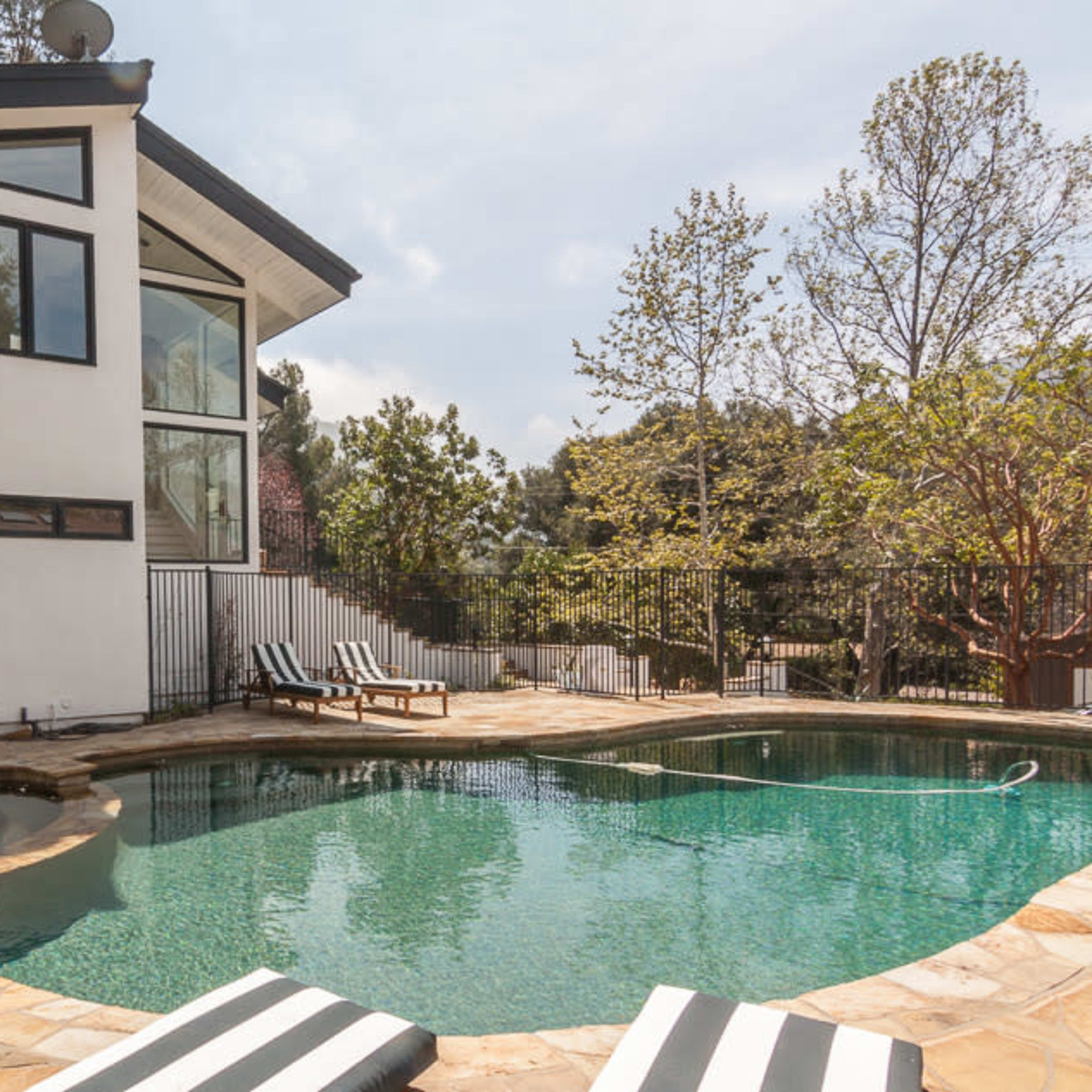 A modern house with large windows overlooking a stone patio and a rectangular swimming pool surrounded by trees.