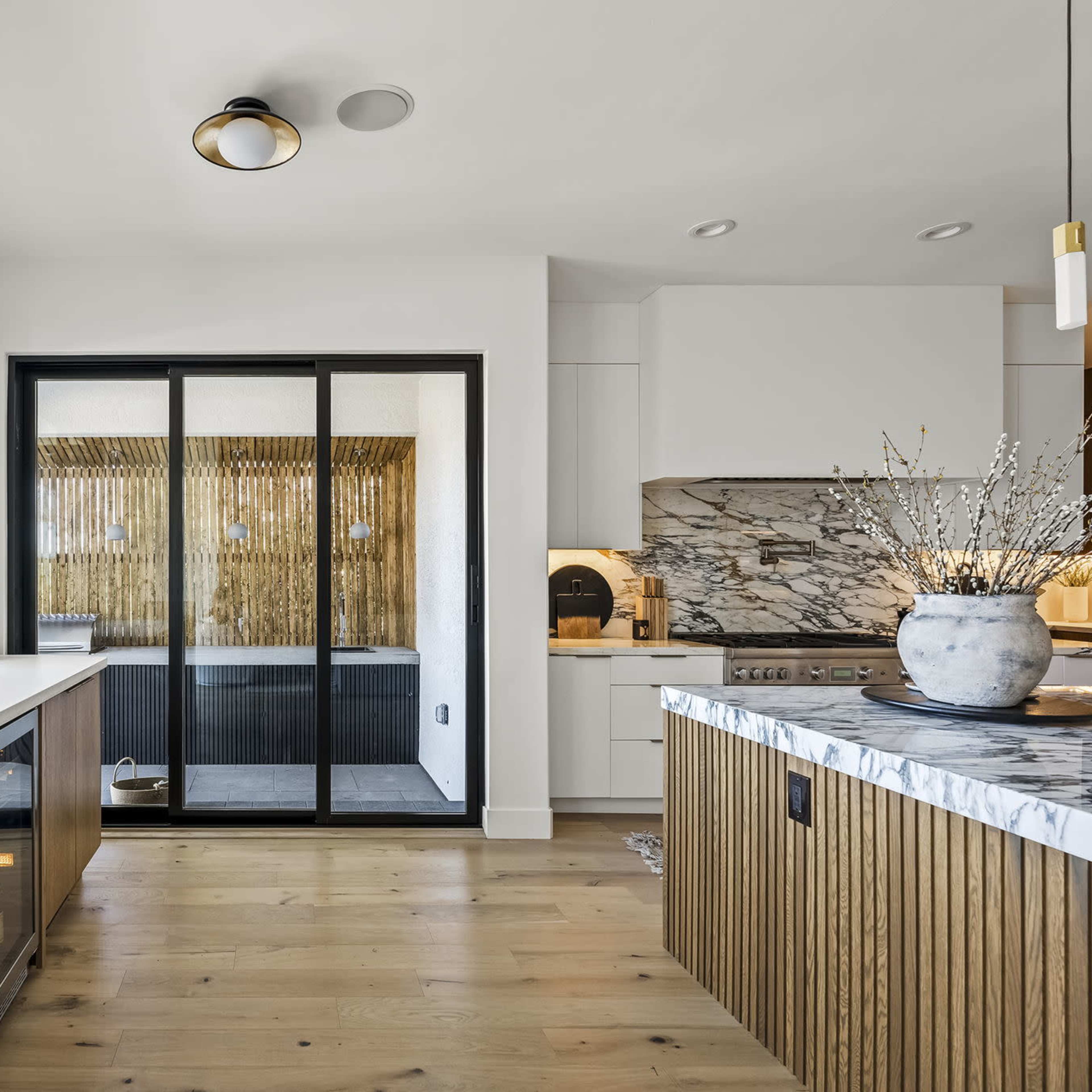 A modern kitchen features wooden cabinetry, a marble-topped island, and large windows that provide natural light.