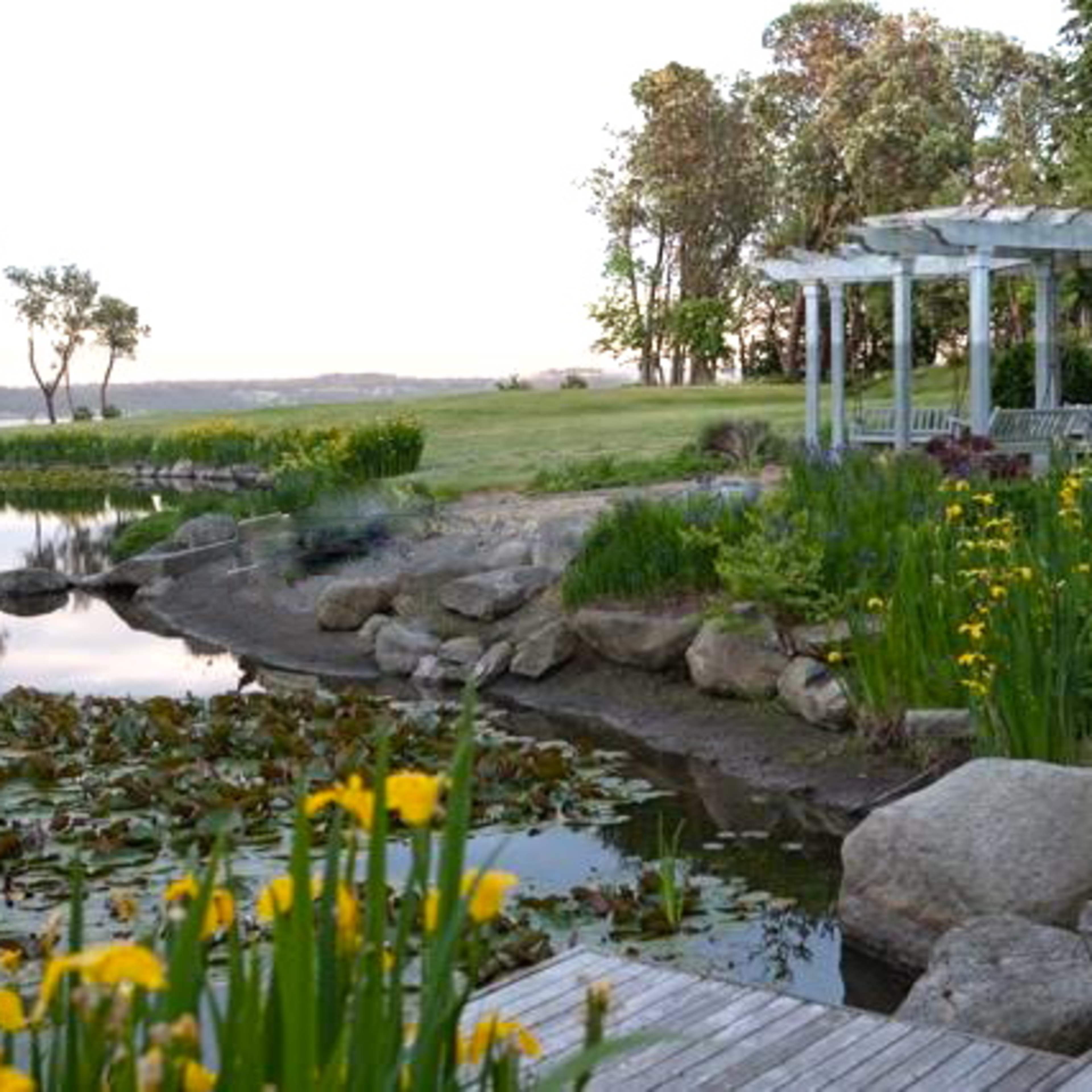 A tranquil lakeside scene features a wooden deck, vibrant yellow flowers, and a pergola near the water, surrounded by greenery and rocky shoreline.