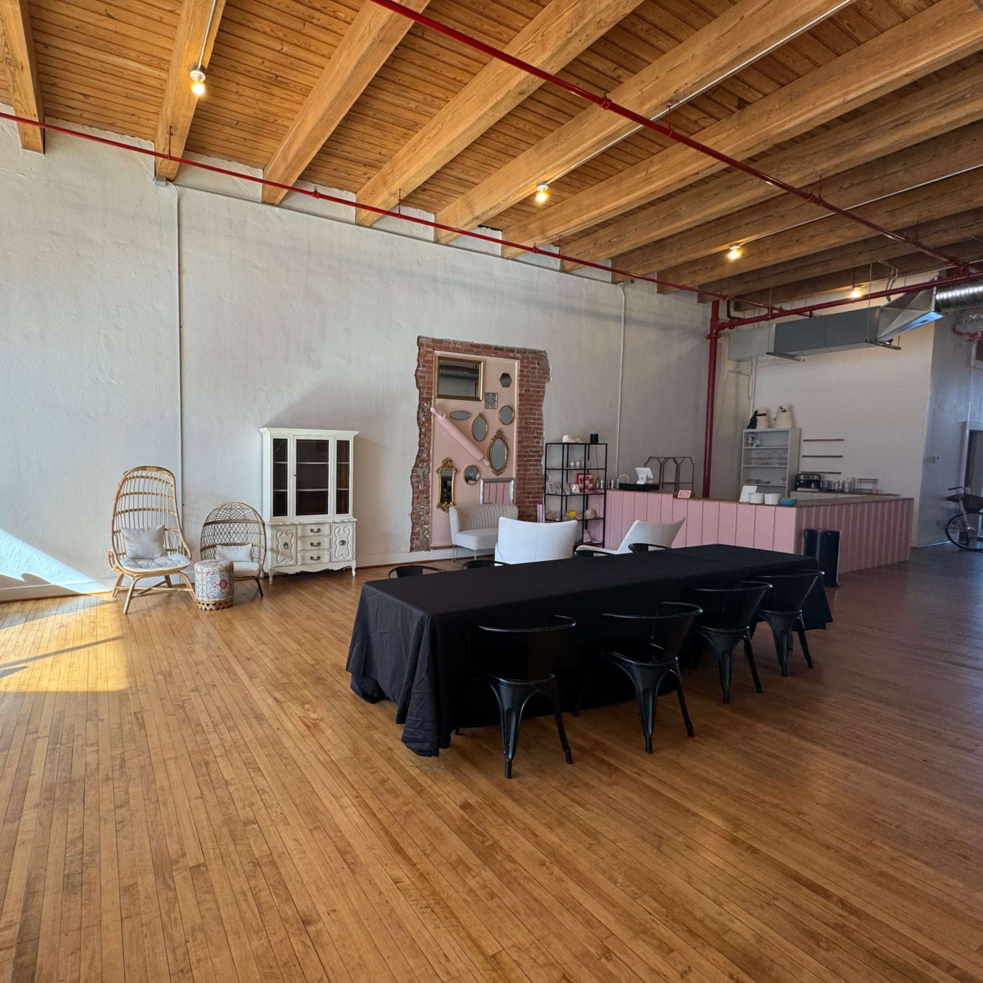 A spacious interior room with wooden floors, exposed beams, a large black table surrounded by chairs, and a mix of vintage and modern furniture.