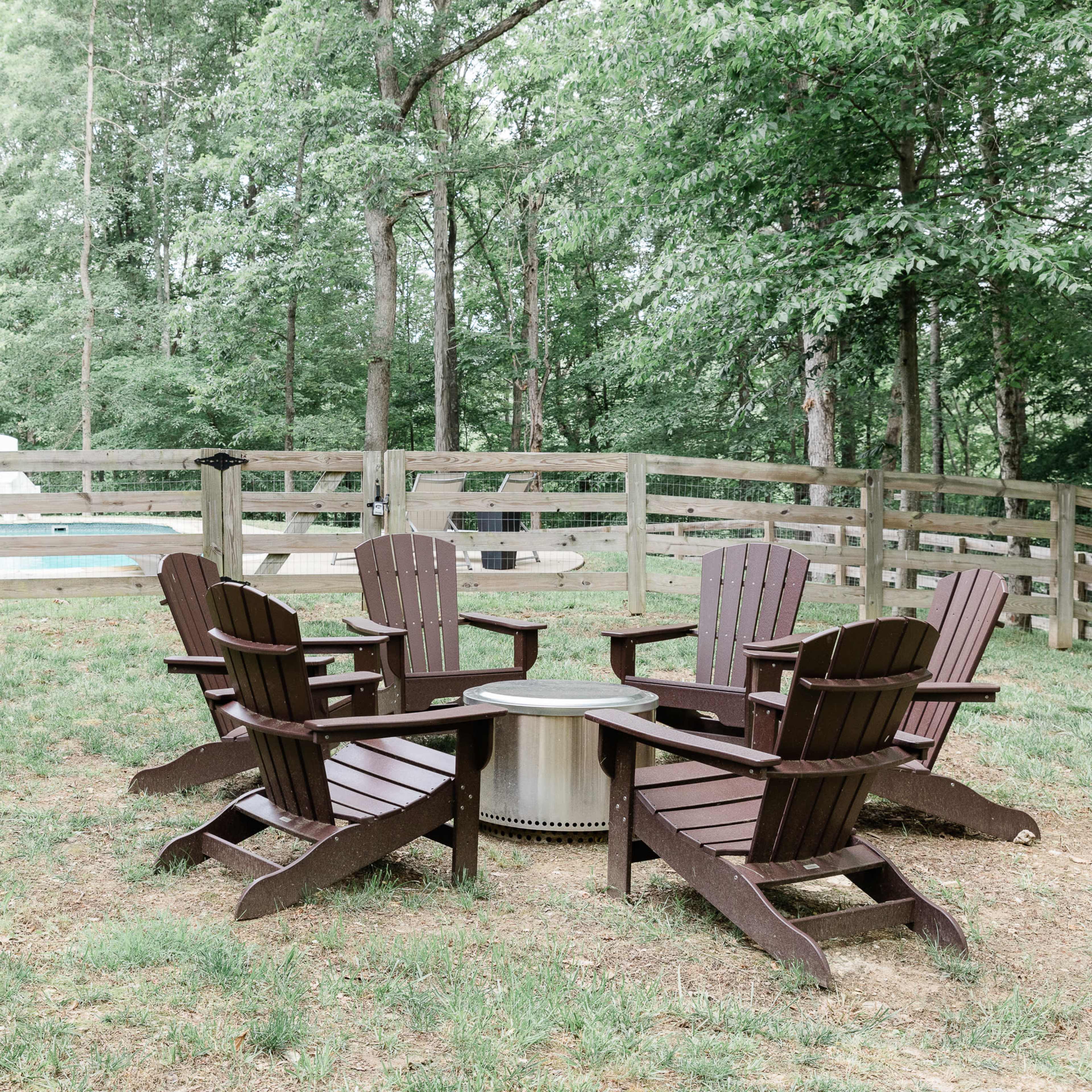 A set of four wooden Adirondack chairs surrounds a fire pit in a grassy area next to a fenced yard with a pool.