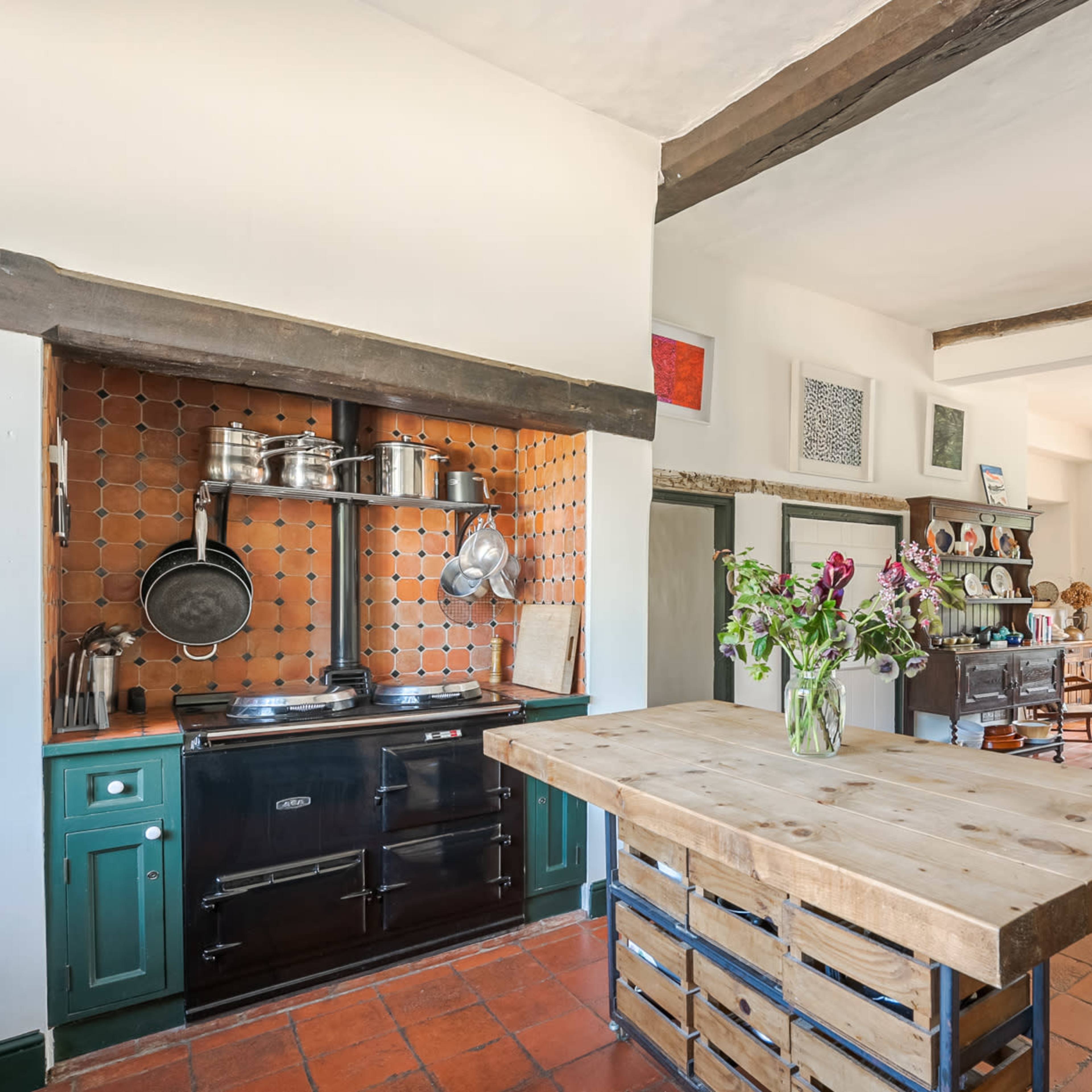 A spacious kitchen featuring a large central wooden island, a black stove with metal cookware, and a dining area with a long table and large windows.