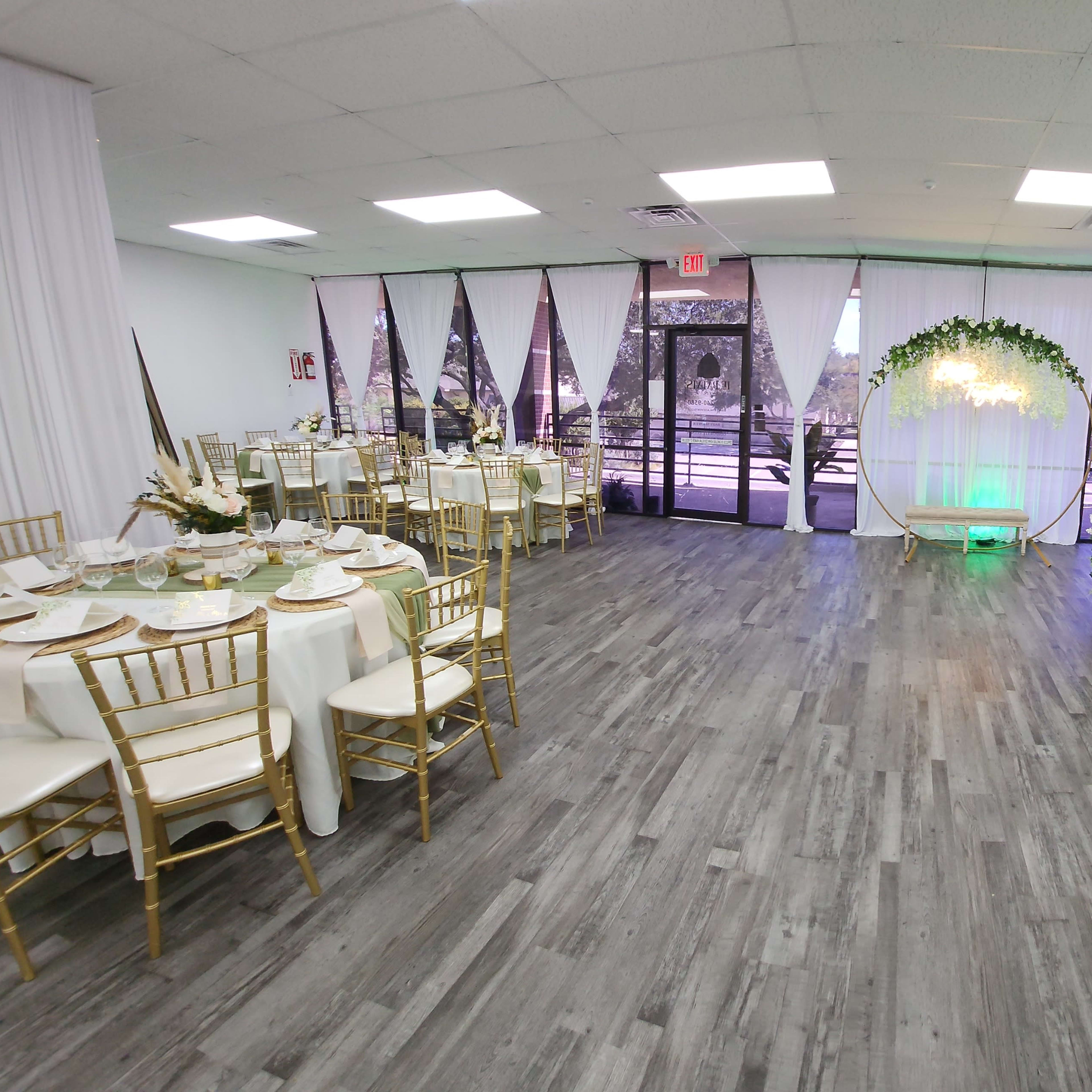 A spacious event venue features elegantly set tables on one side and a decorative backdrop with greenery and lights on the other.