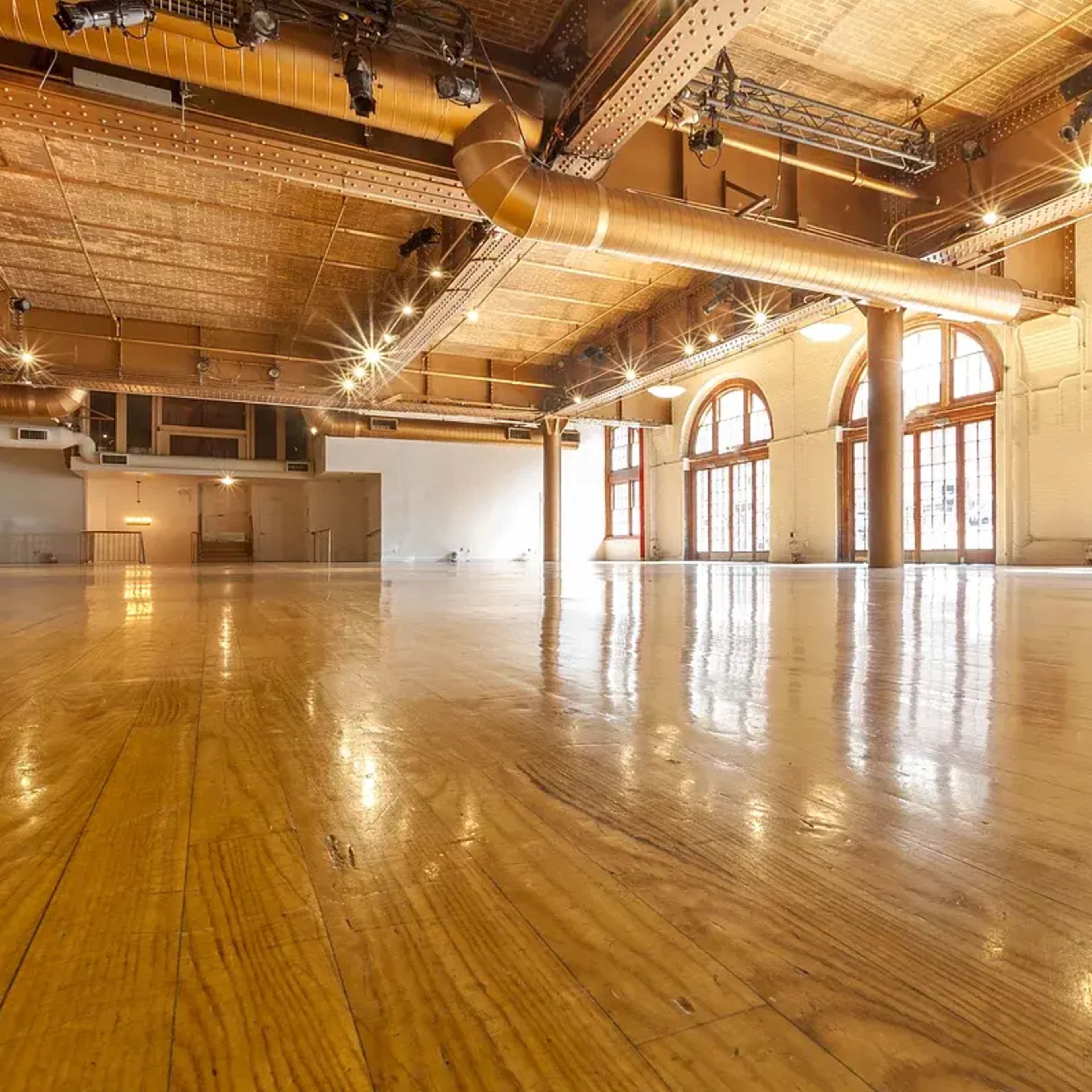 A spacious, empty room with polished wooden floors and large windows allowing natural light.