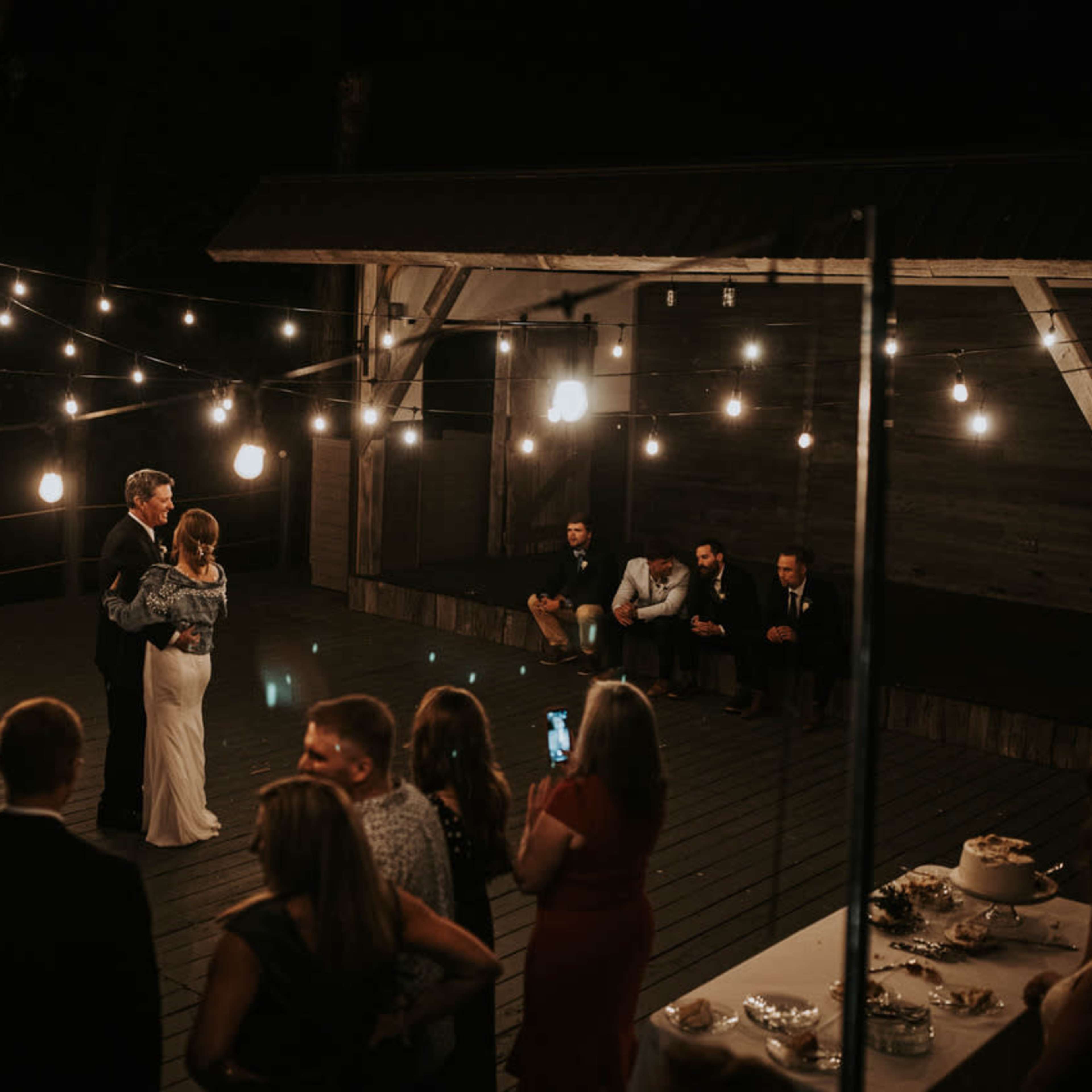 A couple dances under string lights on a wooden deck while guests watch and take photos.