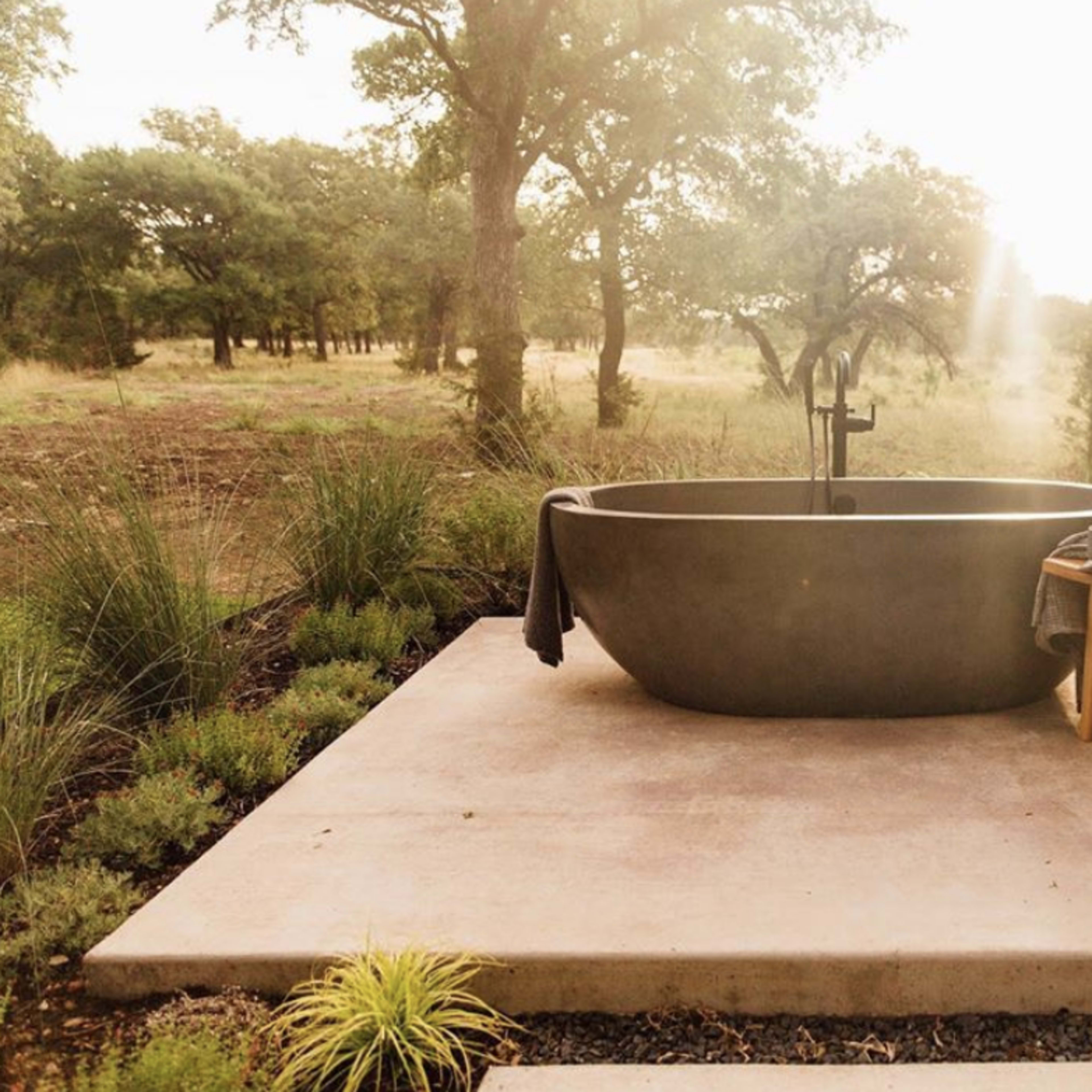 A modern freestanding bathtub sits on a concrete platform surrounded by greenery in a natural setting.