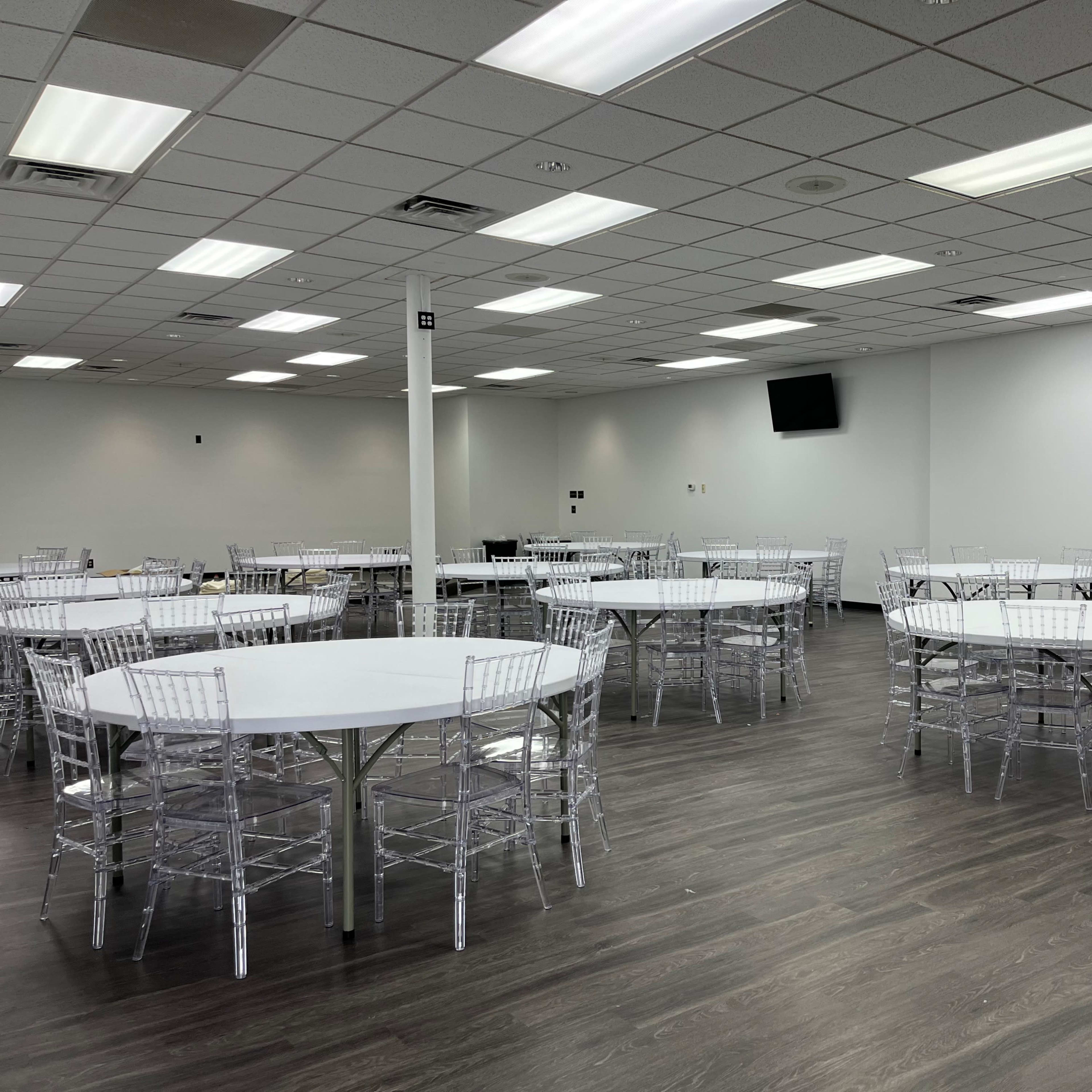 A spacious room with multiple round tables and clear acrylic chairs arranged in a neat layout.