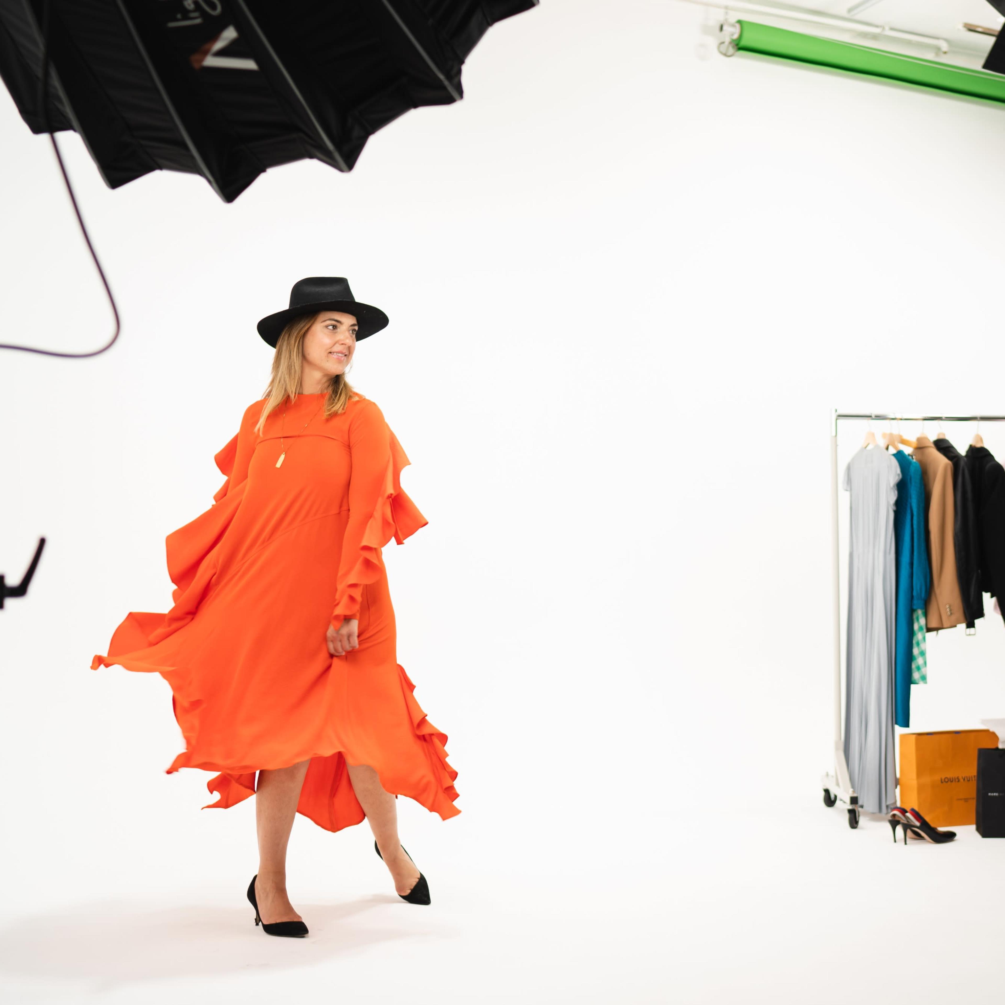 A model wearing an orange dress and a black hat walks on a white backdrop, with a clothing rack and photography lights in the background.