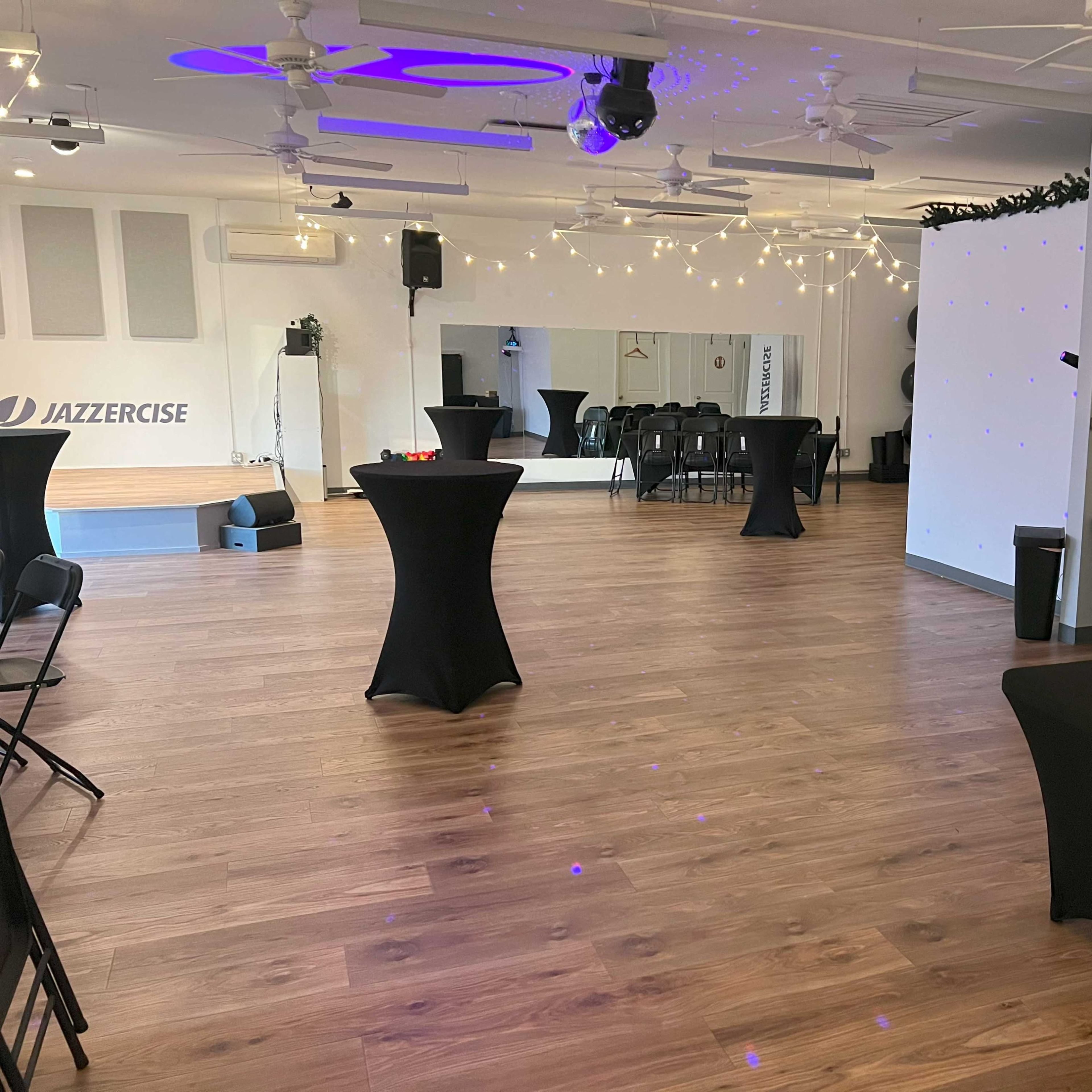 A spacious, well-lit event room with several black tables and chairs arranged for a gathering, featuring string lights overhead and a mirrored wall on one side.