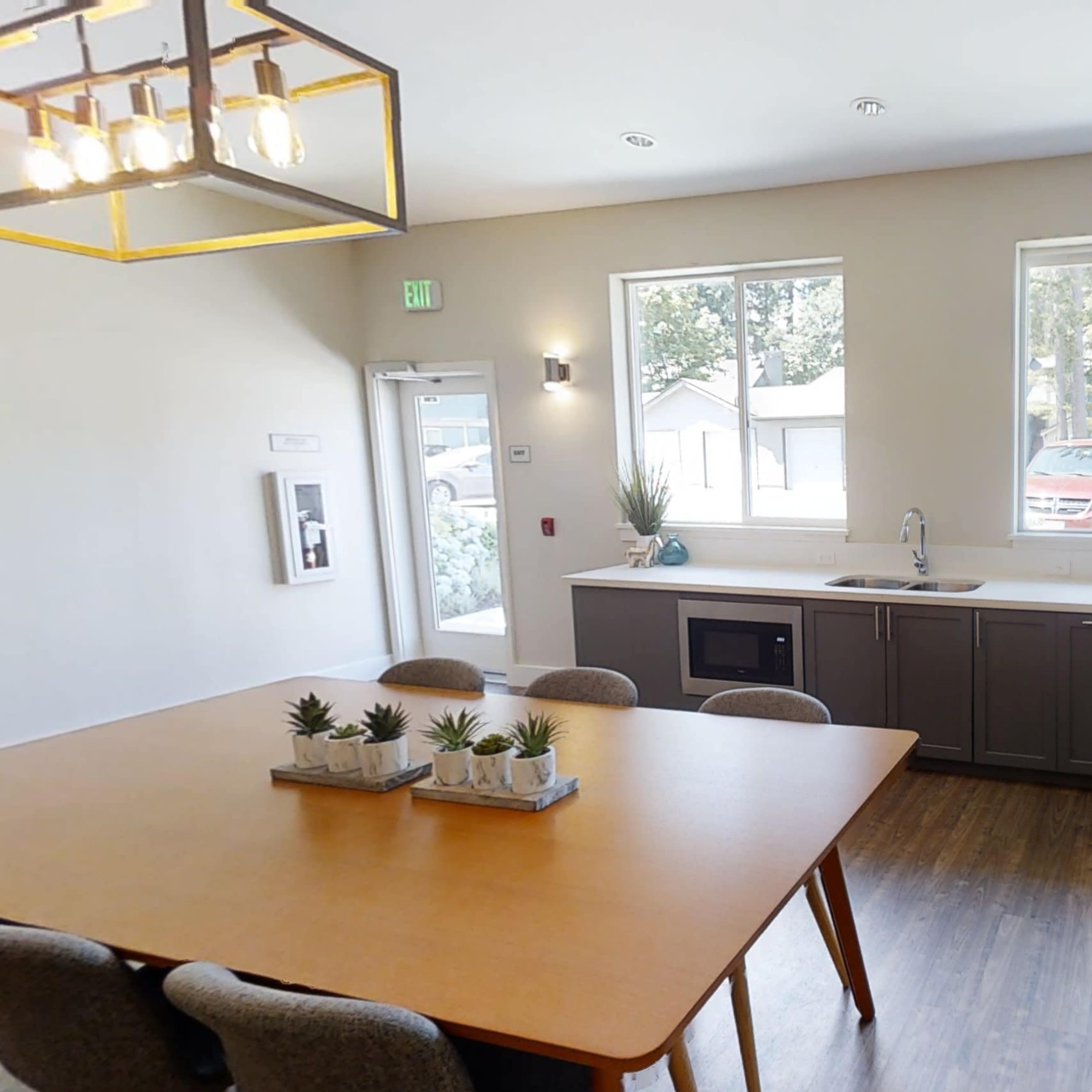 A modern kitchen area with a large wooden table, a stainless steel refrigerator, and windows providing natural light.