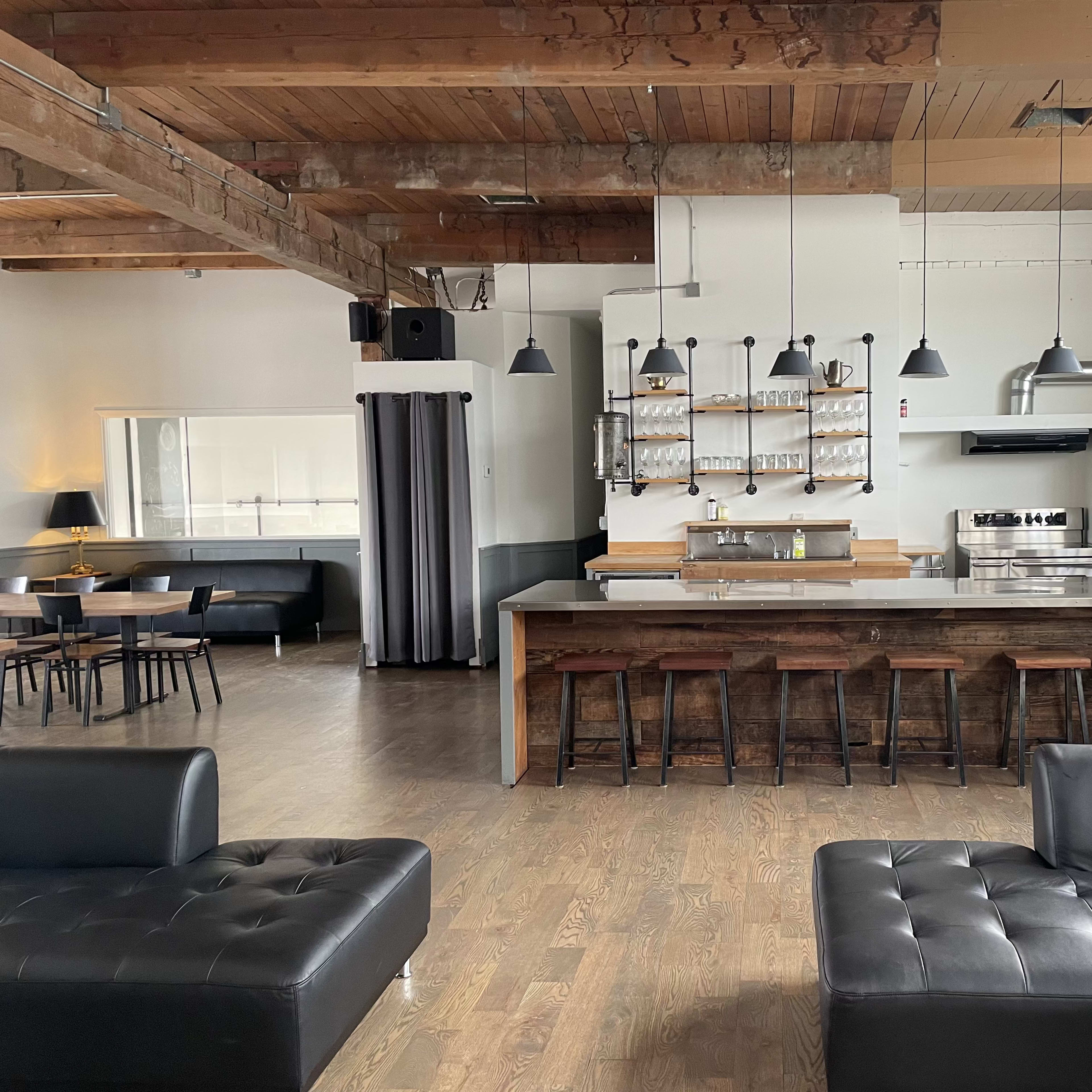 A modern kitchen and dining area featuring wooden beams, a large countertop with bar stools, and seating arrangements with dark couches.