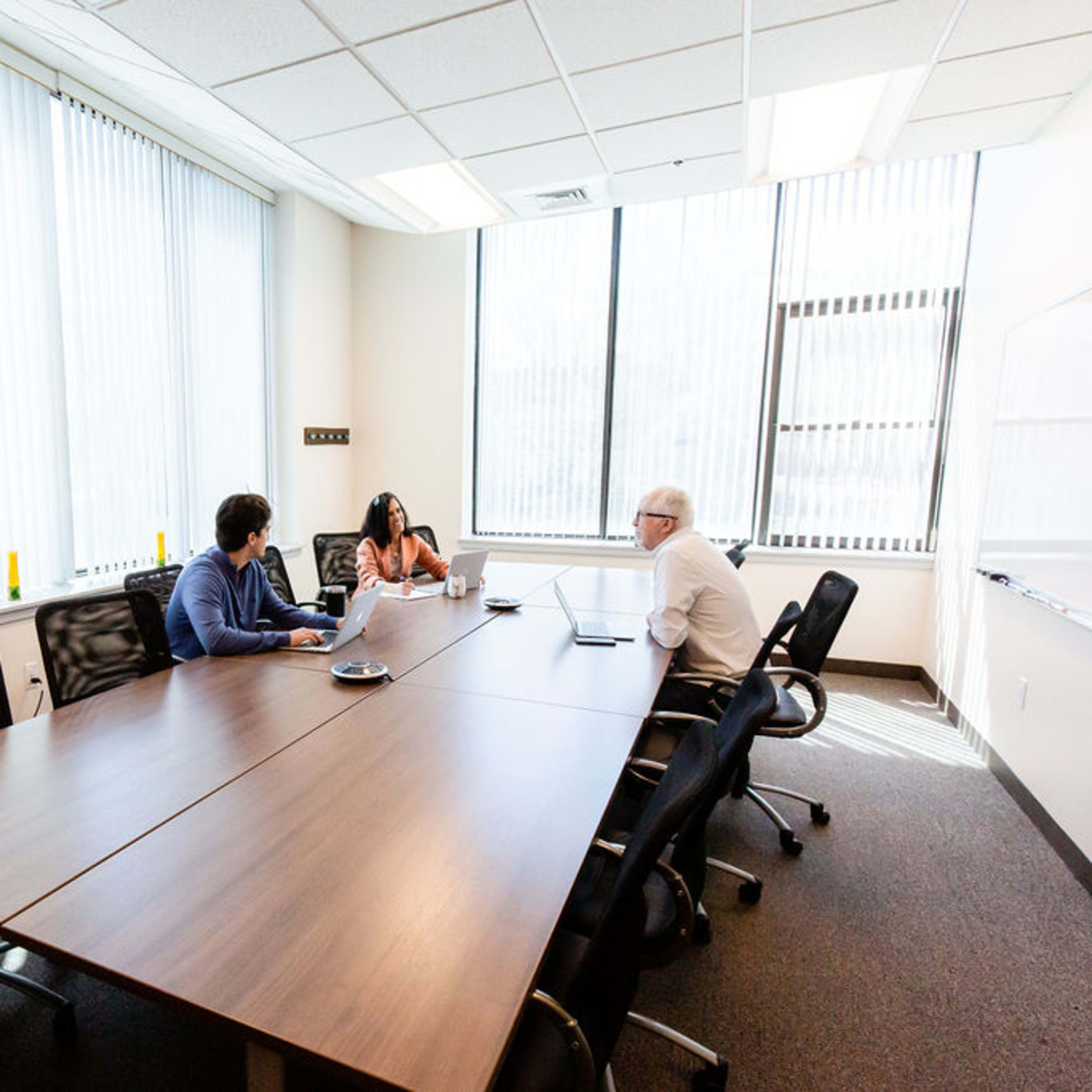 Three people are seated around a conference table in a well-lit meeting room with large windows and blinds.