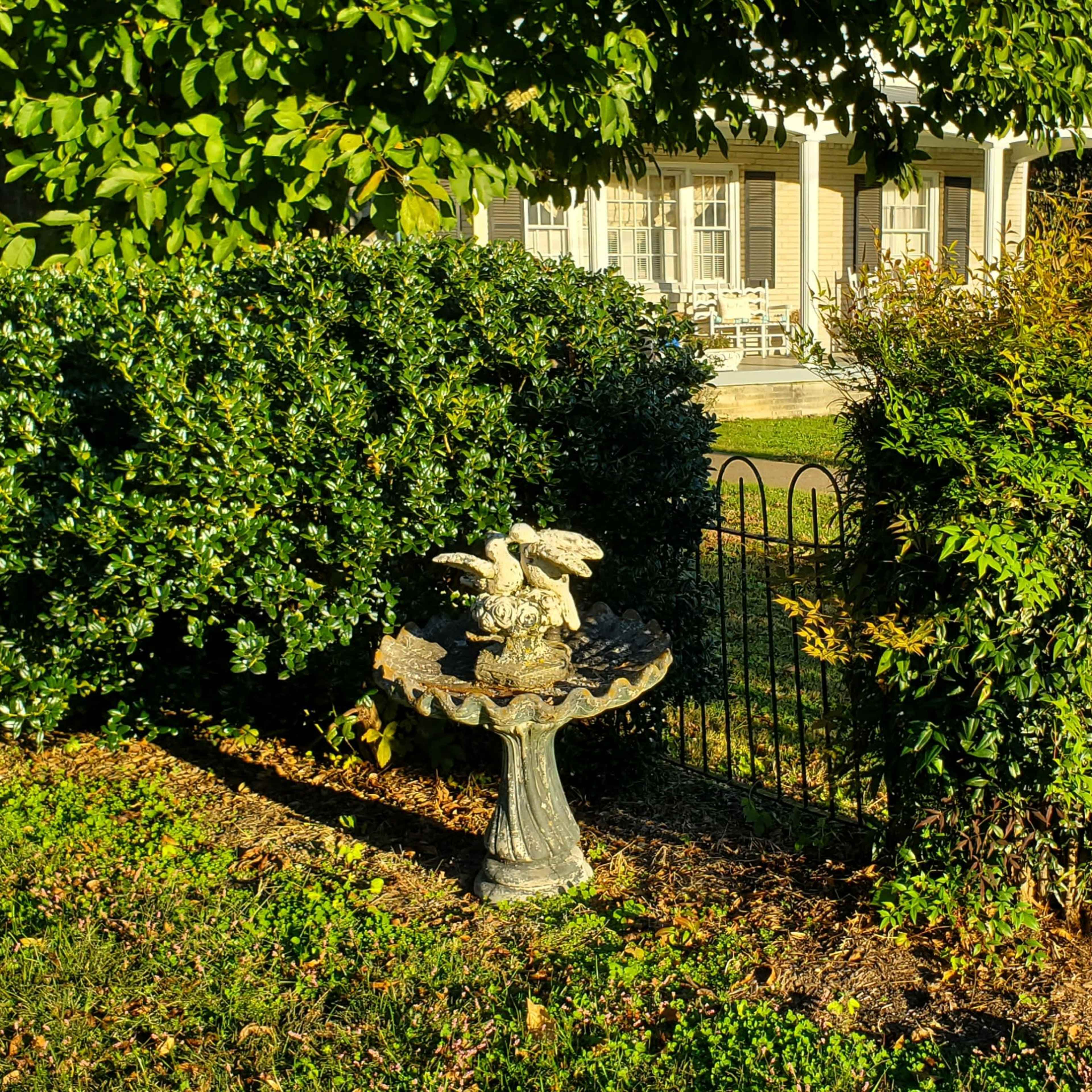 A birdbath with a figurine is surrounded by greenery near a house with a porch.