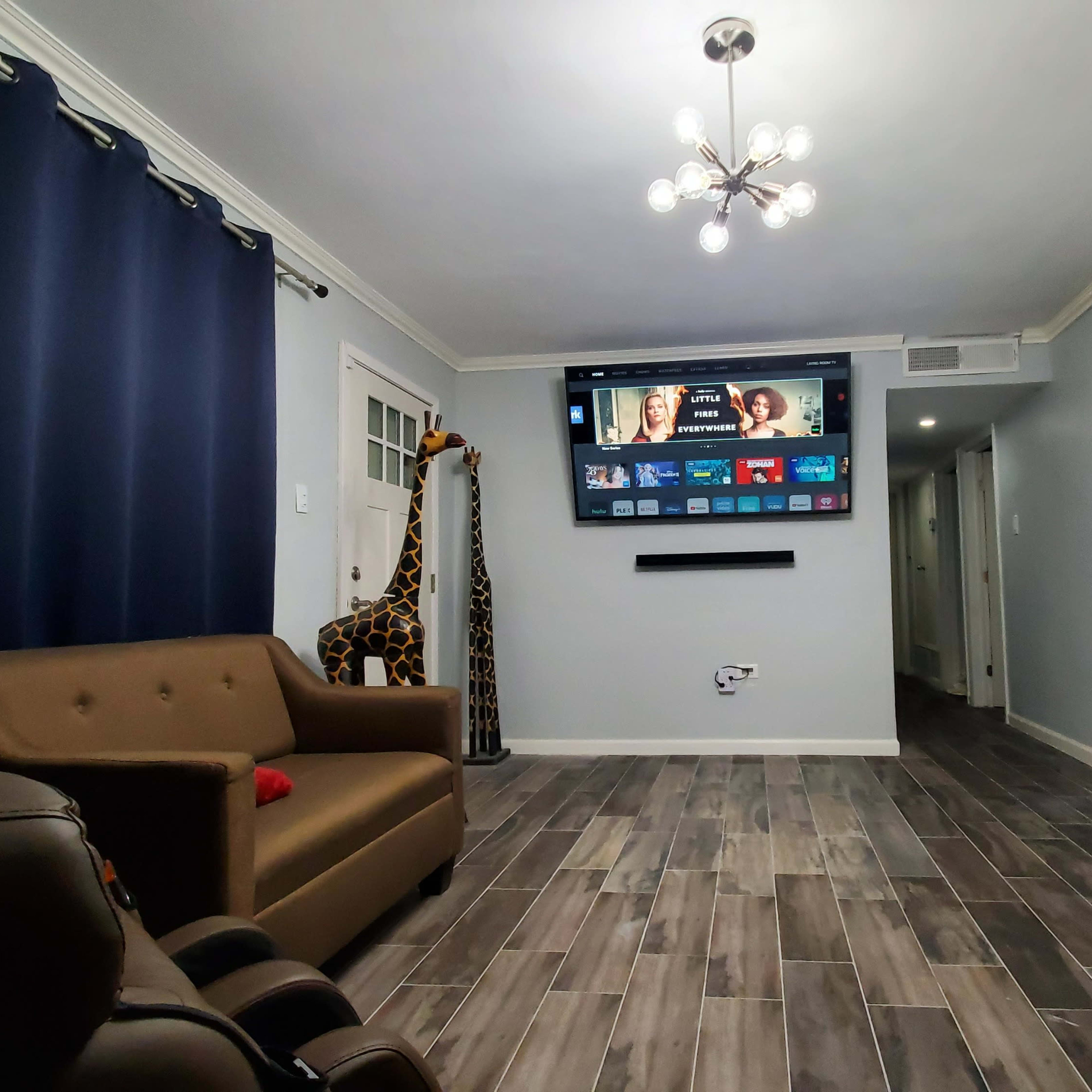 A living room featuring a brown sofa, a tall giraffe decoration, a mounted TV displaying a screen interface, and walls painted in light colors.