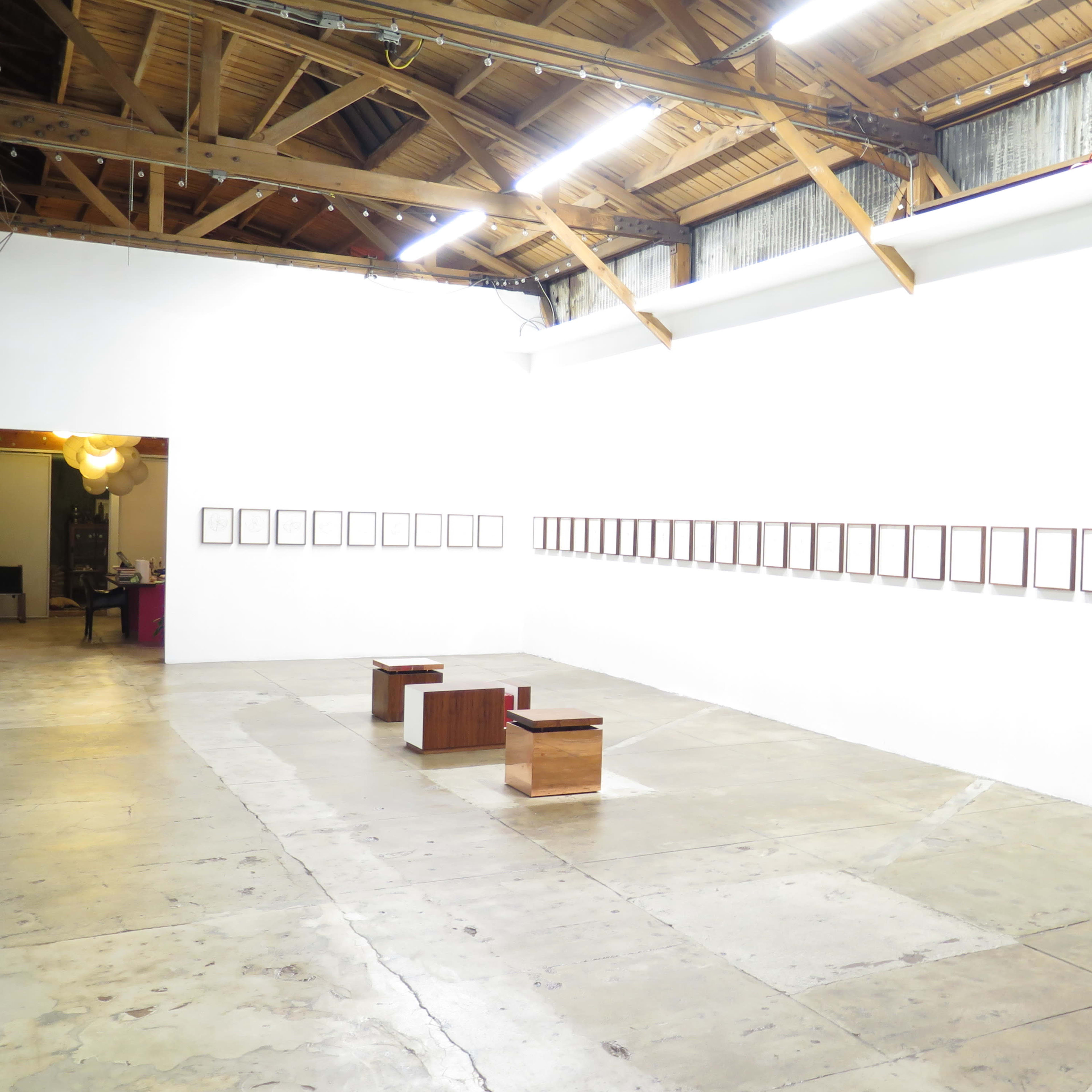 A gallery interior featuring a series of framed artworks displayed on a white wall, with wooden benches positioned in the center of the room.