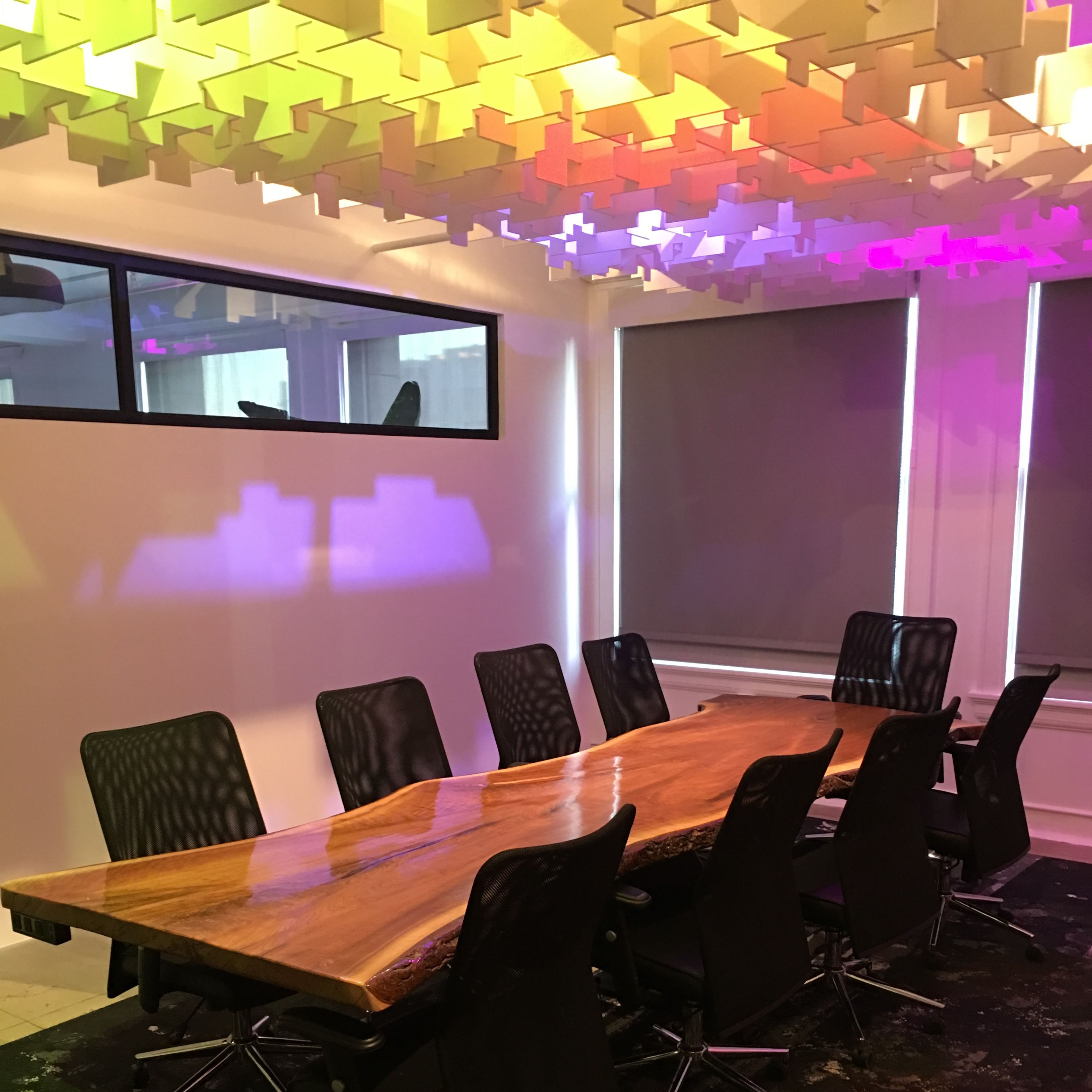 A modern conference room features a large wooden table surrounded by black chairs, with colorful lights illuminating the ceiling and walls.