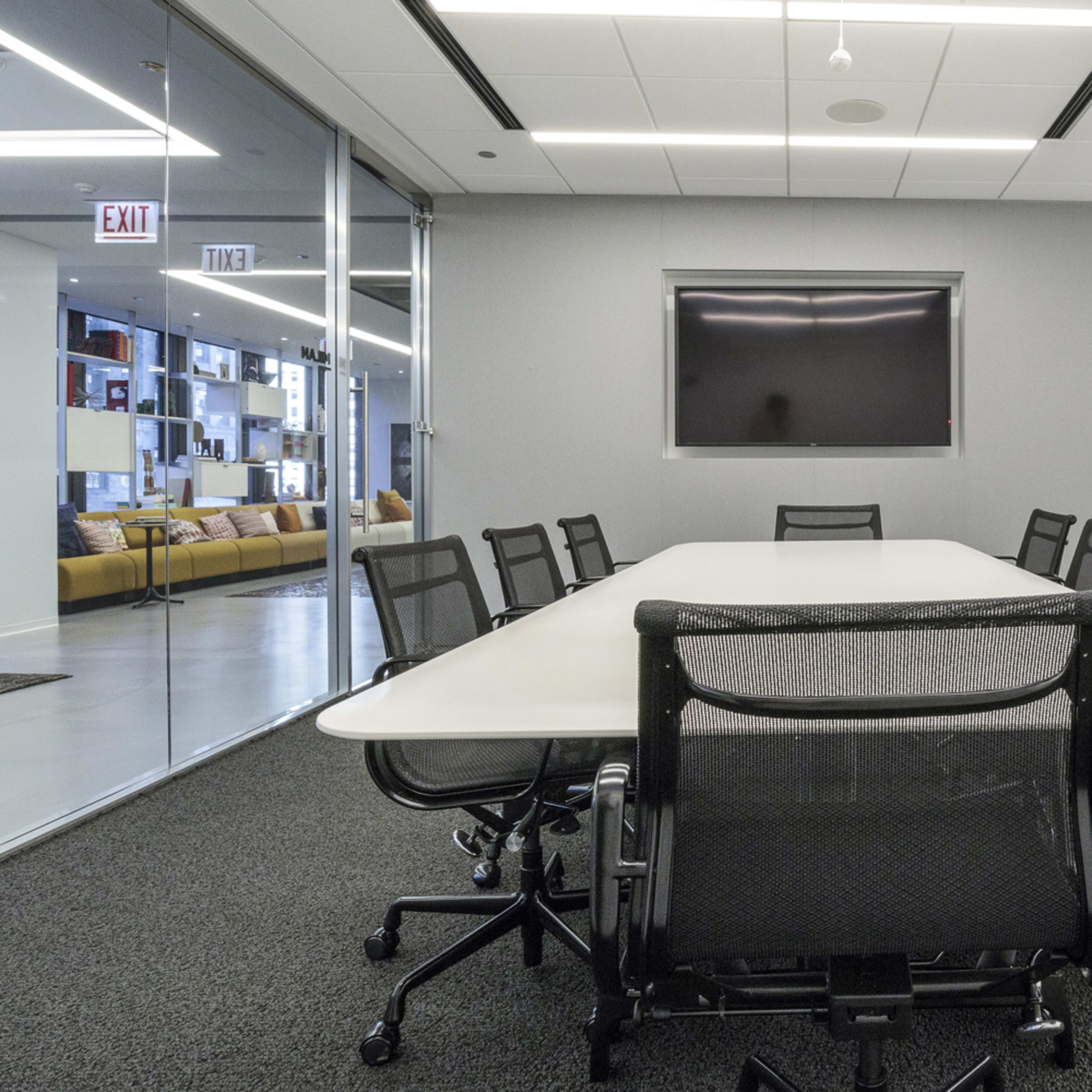 A modern conference room features a long rectangular table surrounded by ergonomic chairs and a wall-mounted screen.