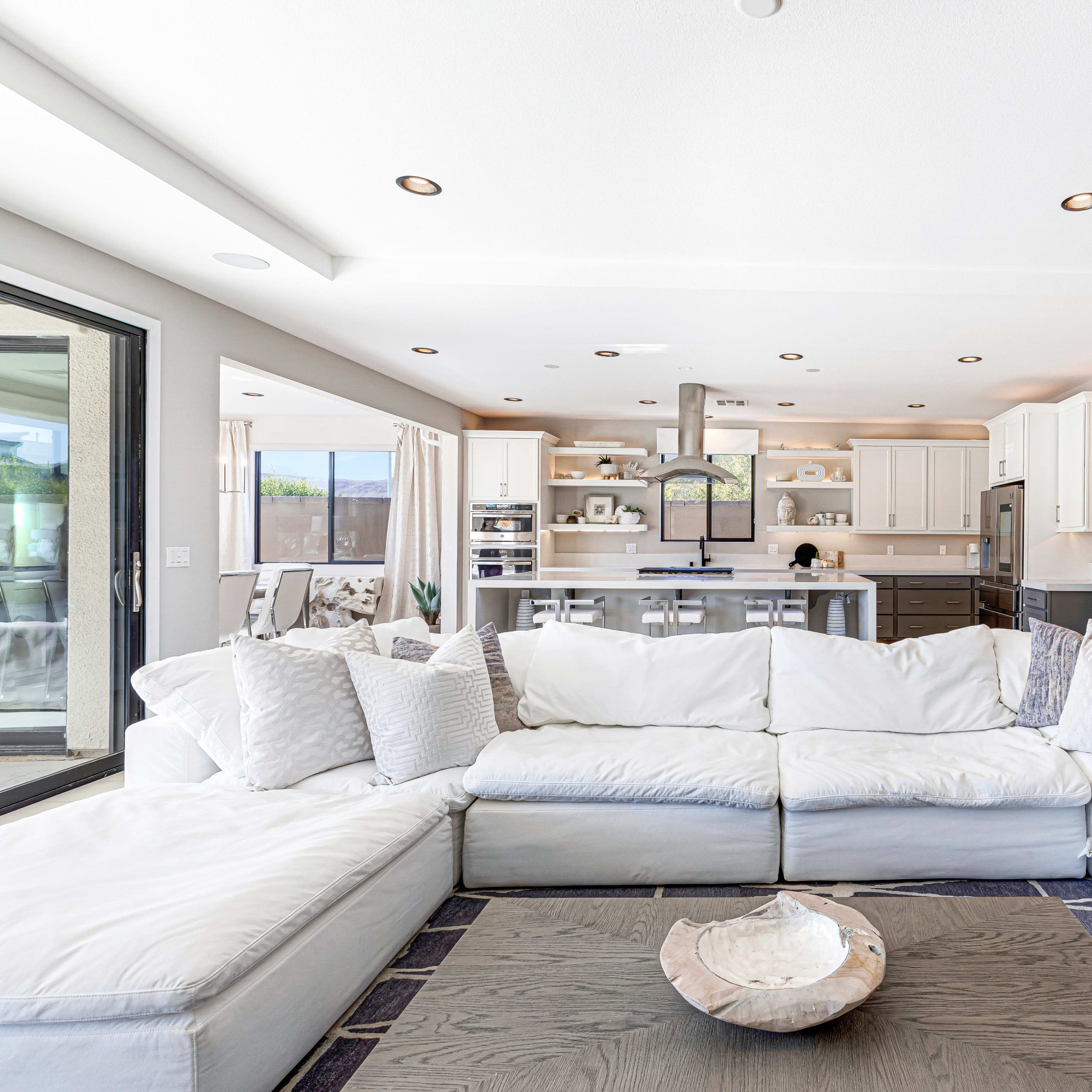 A spacious living room features a large white sectional sofa facing an open kitchen with modern appliances and natural light.