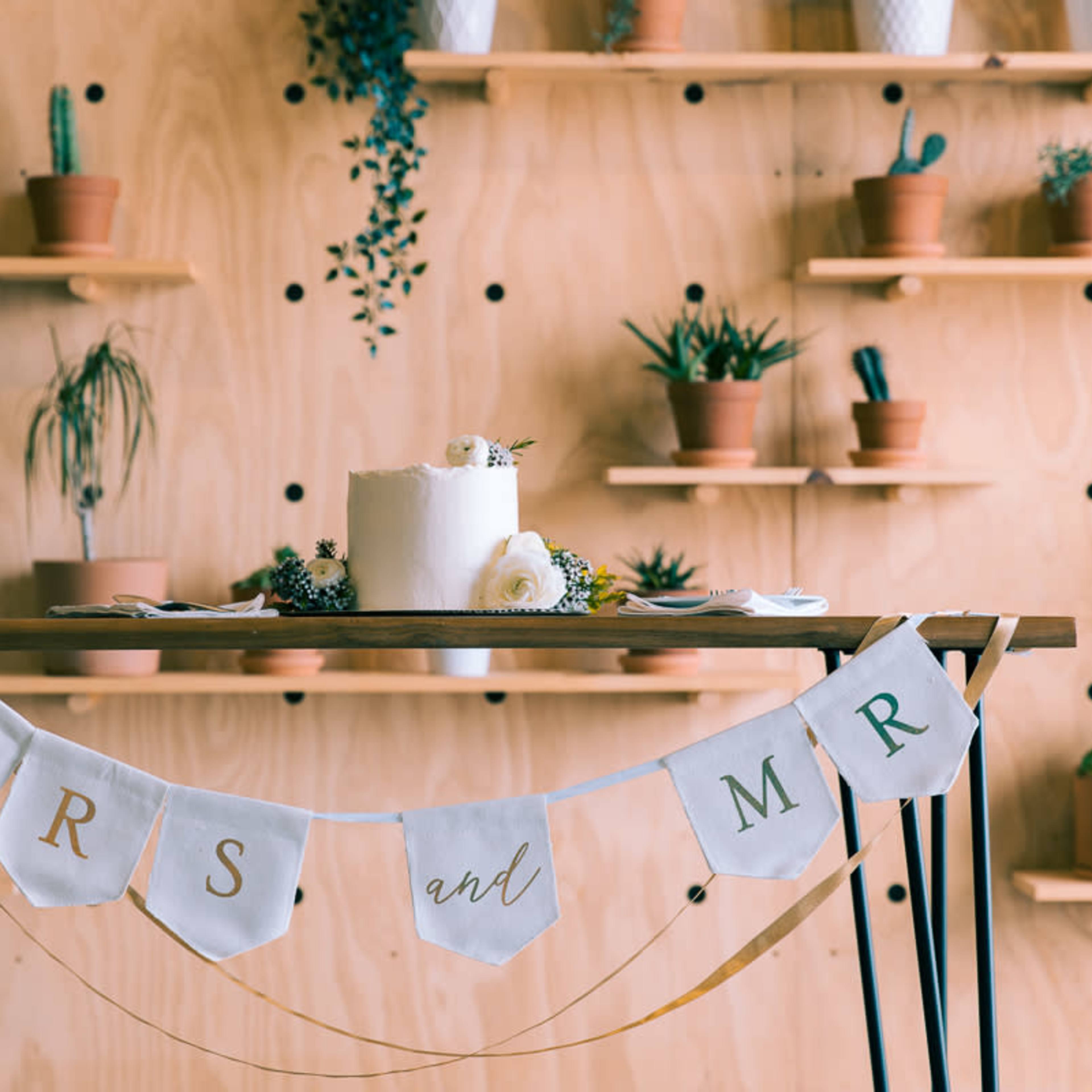 A table decorated with a white cake and a banner reading "MRS and MR," set against a backdrop of potted plants on wooden shelves.