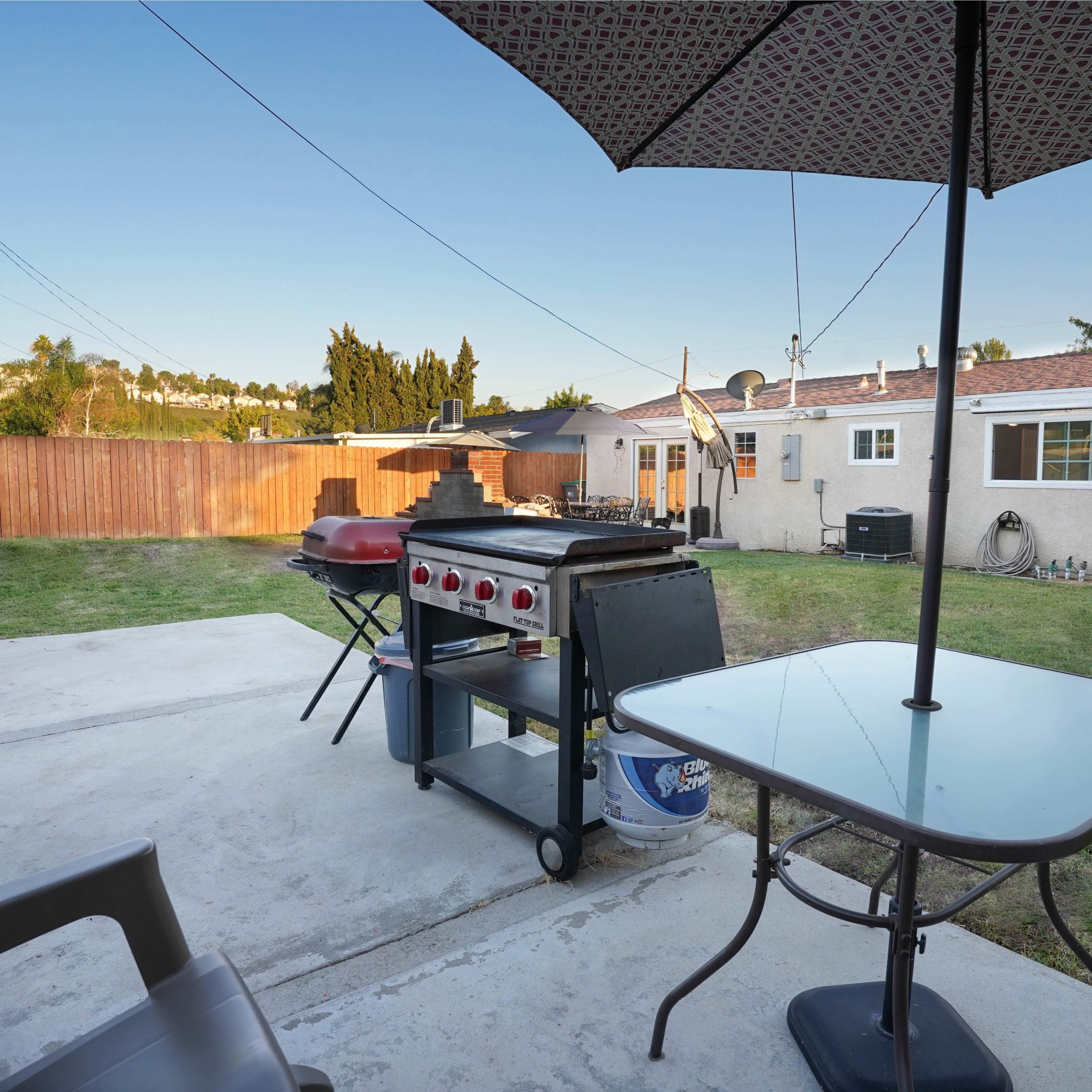 A backyard with a grill on a concrete patio, a small table and chair, and houses in the background.