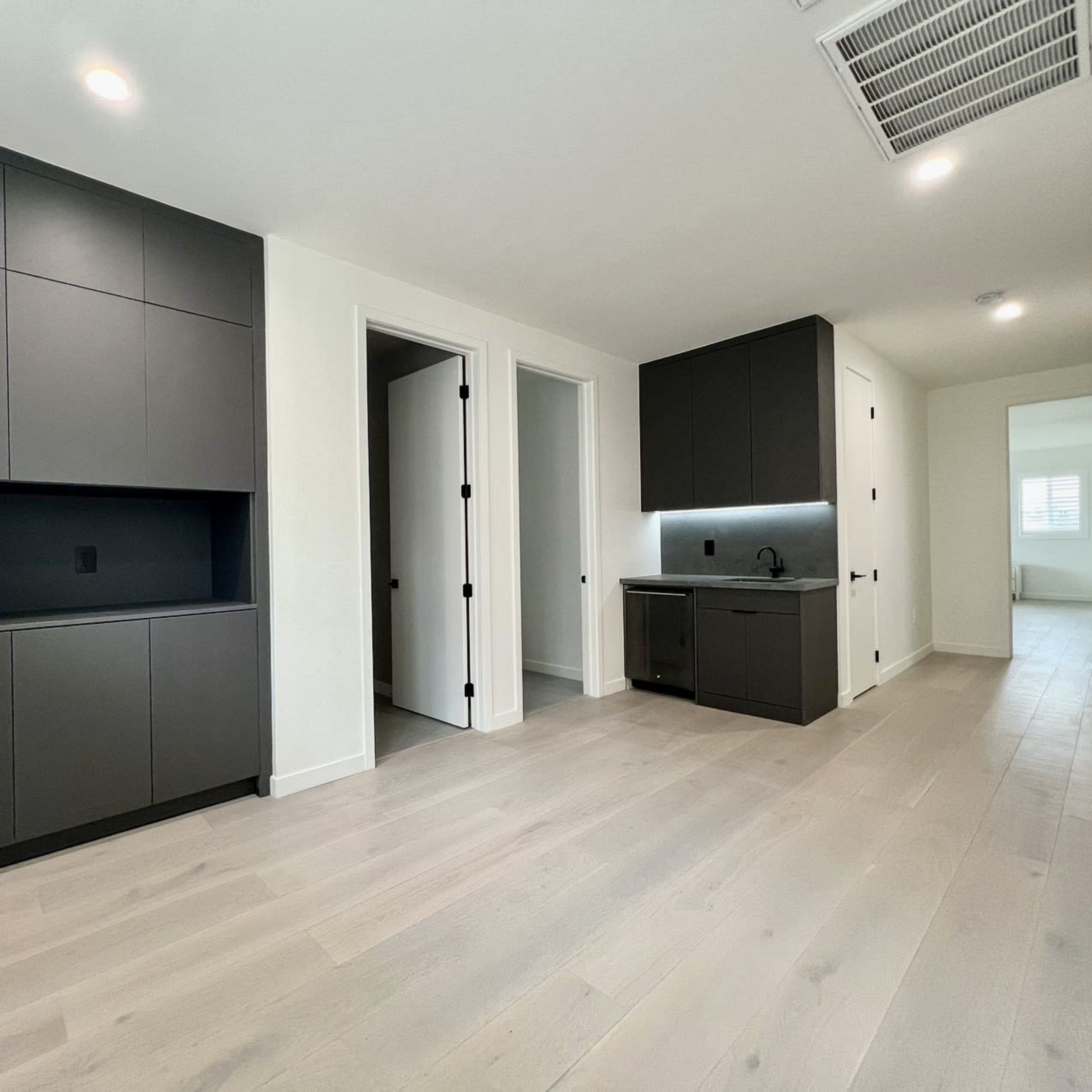 A modern, minimalist interior space with light hardwood floors, dark cabinetry, and two doorways leading to separate rooms.