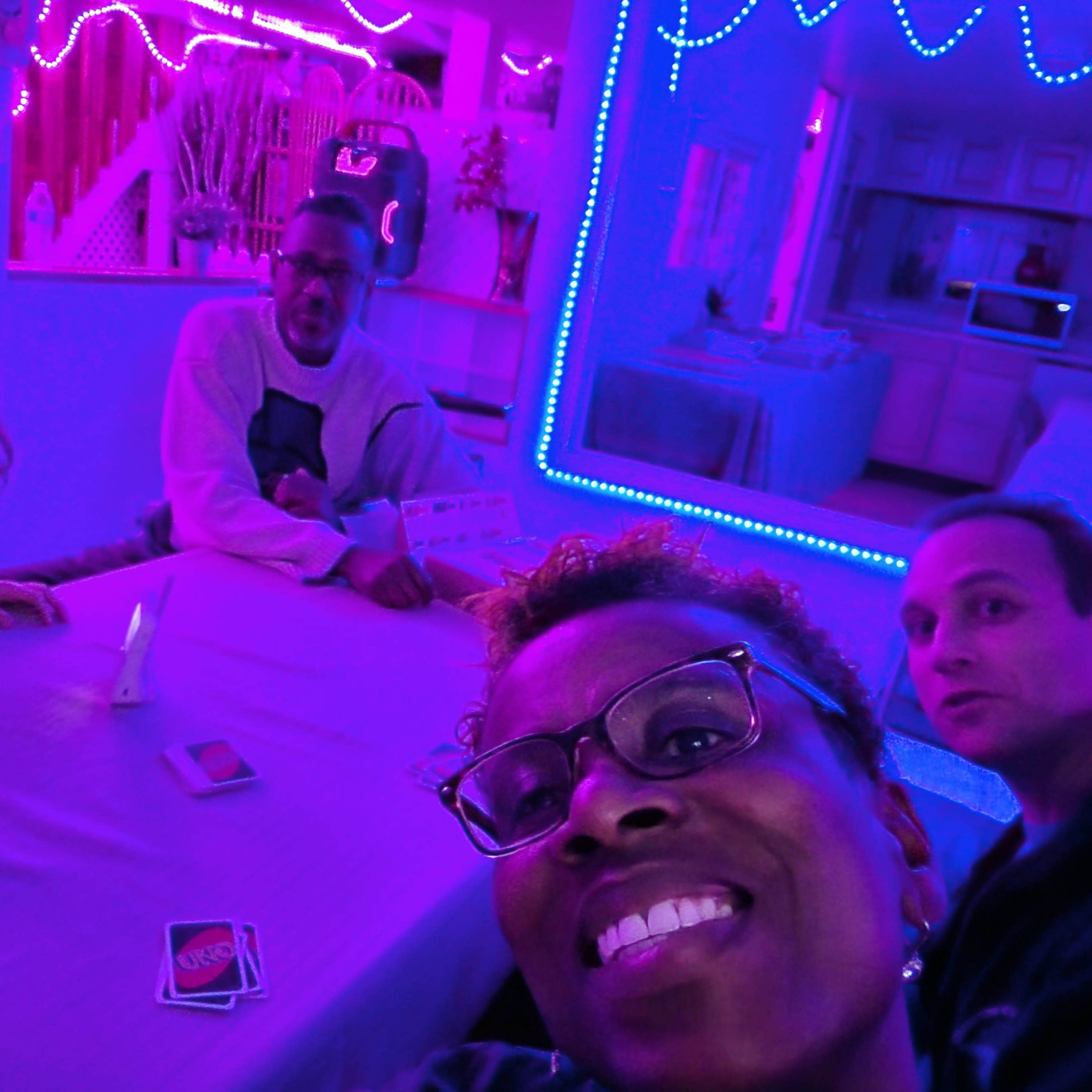 A group of four people gathers around a table illuminated by colored neon lights in a room.