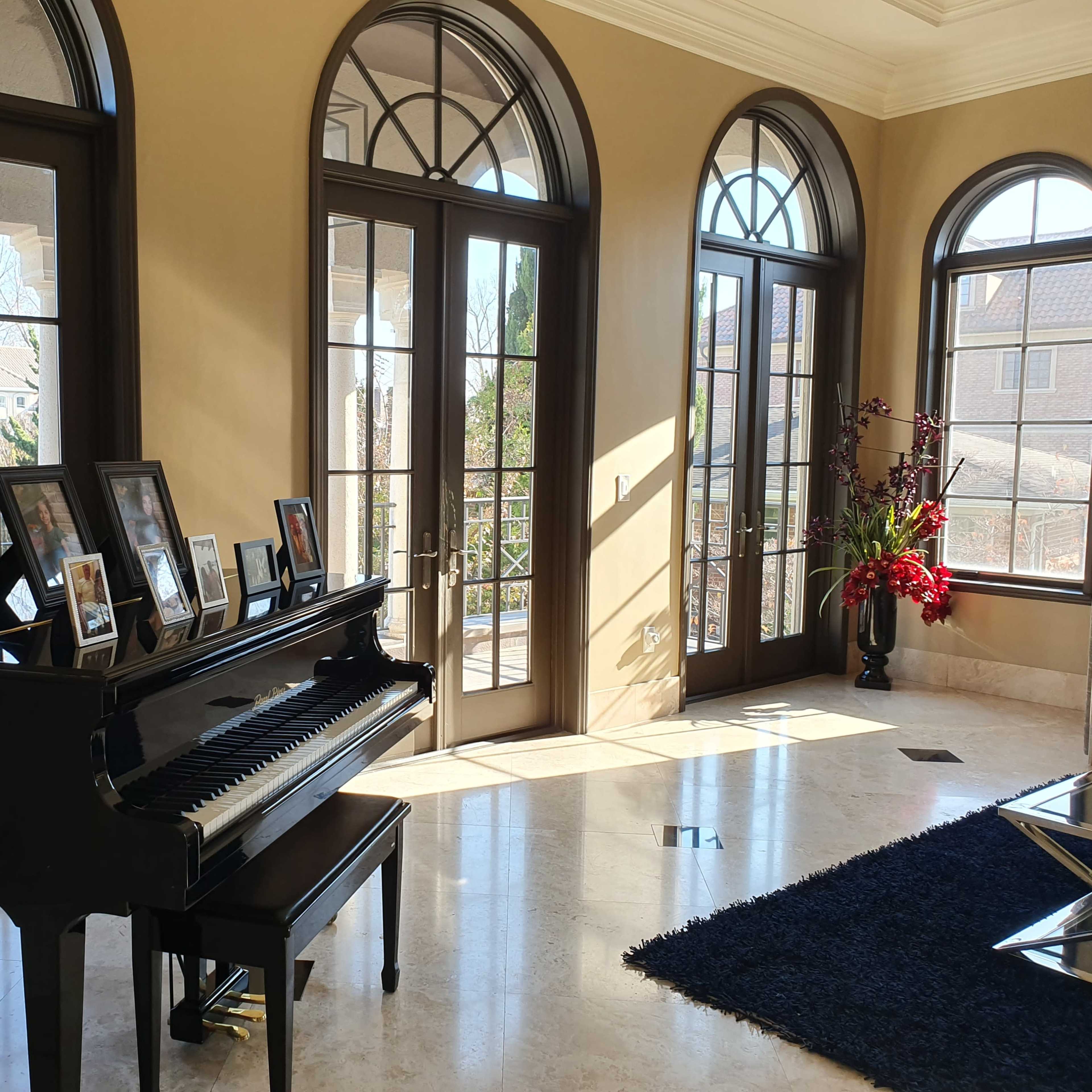A black piano sits beside a wall adorned with photo frames, illuminated by natural light streaming through large arched windows in a spacious room.
