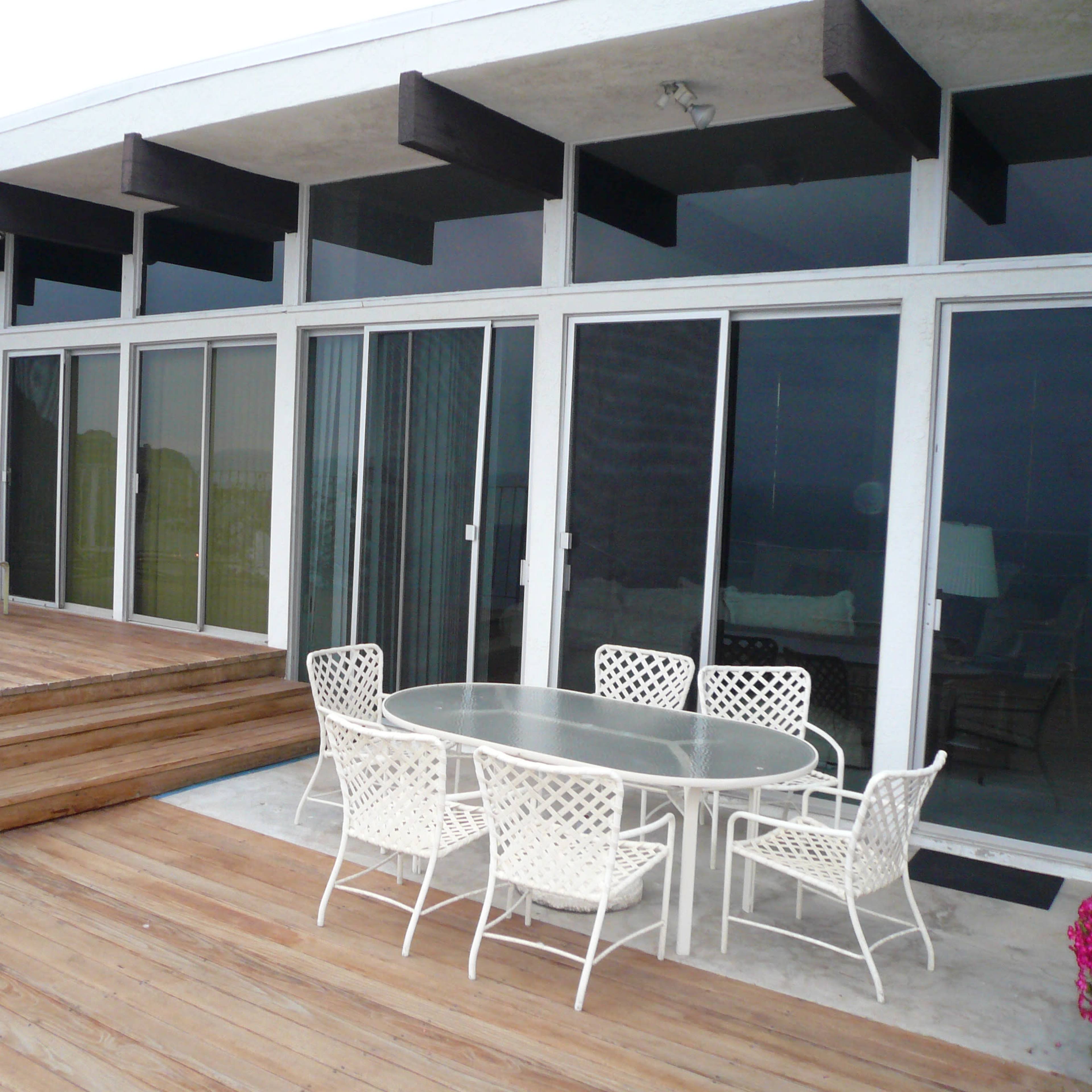 A patio with a glass table and white chairs is situated in front of a row of large windows at a modern building.