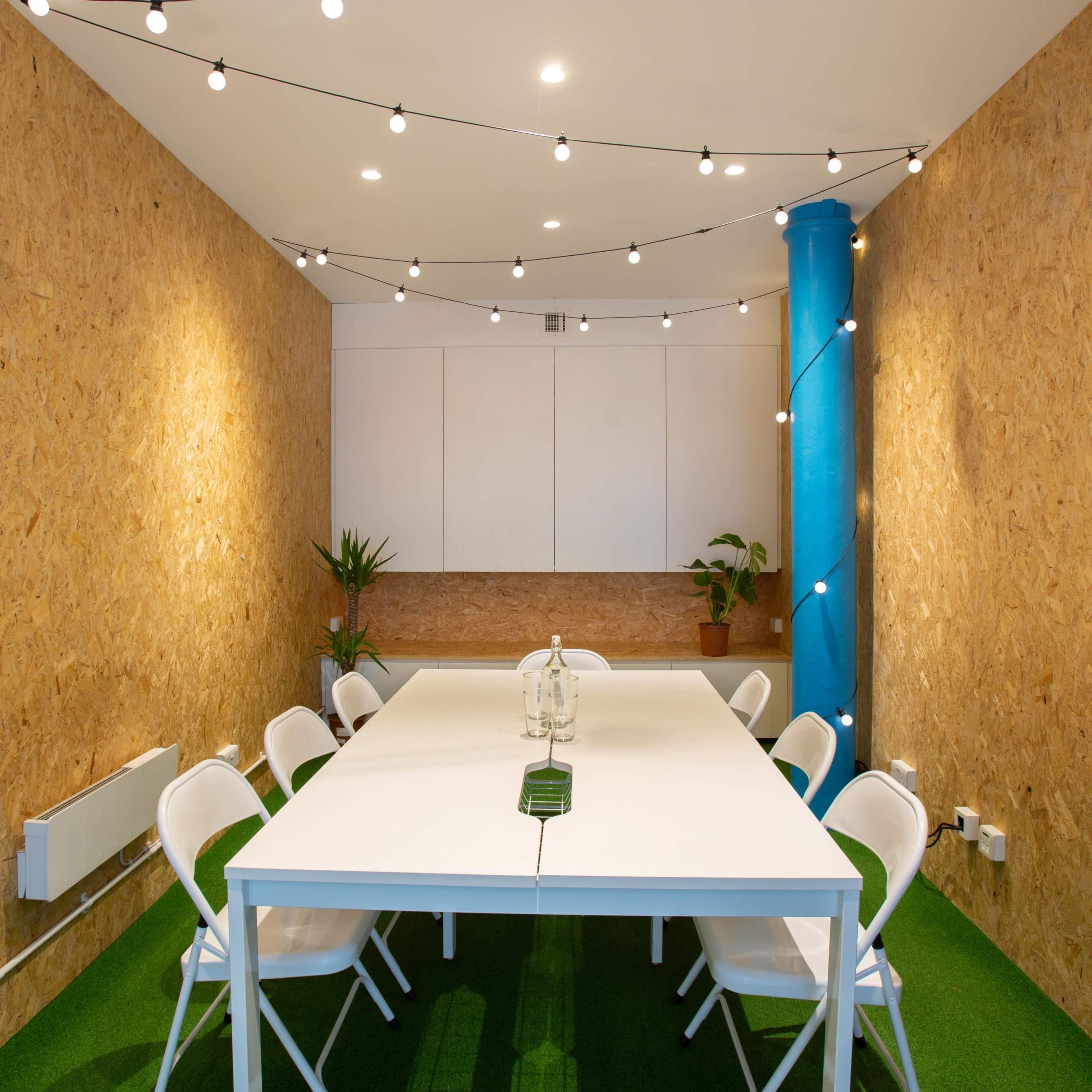 A meeting room features a rectangular white table surrounded by eight white chairs, with wooden walls and a green carpet, illuminated by string lights overhead.