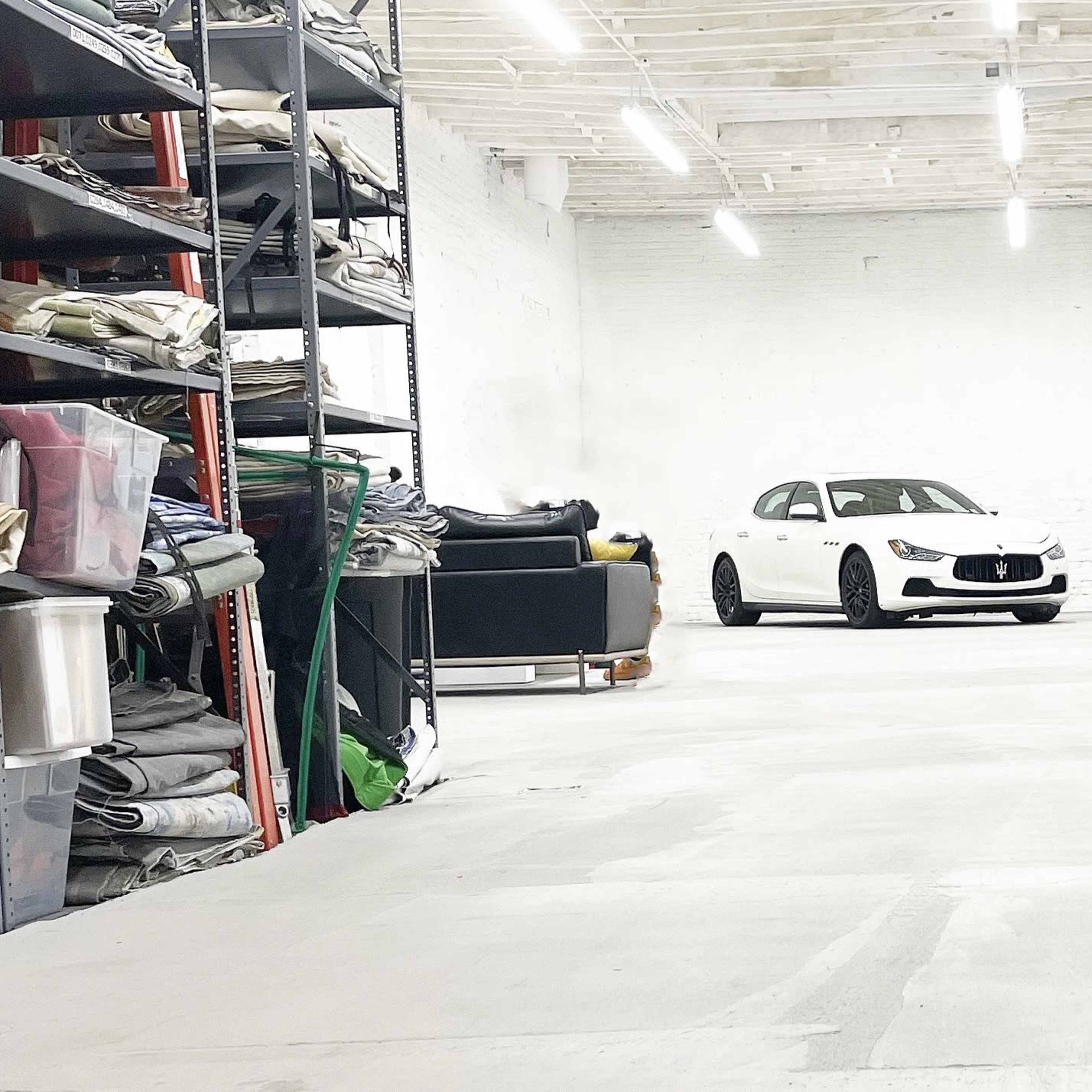 A white car is parked in a spacious, empty warehouse with shelves of materials and equipment lining the walls.