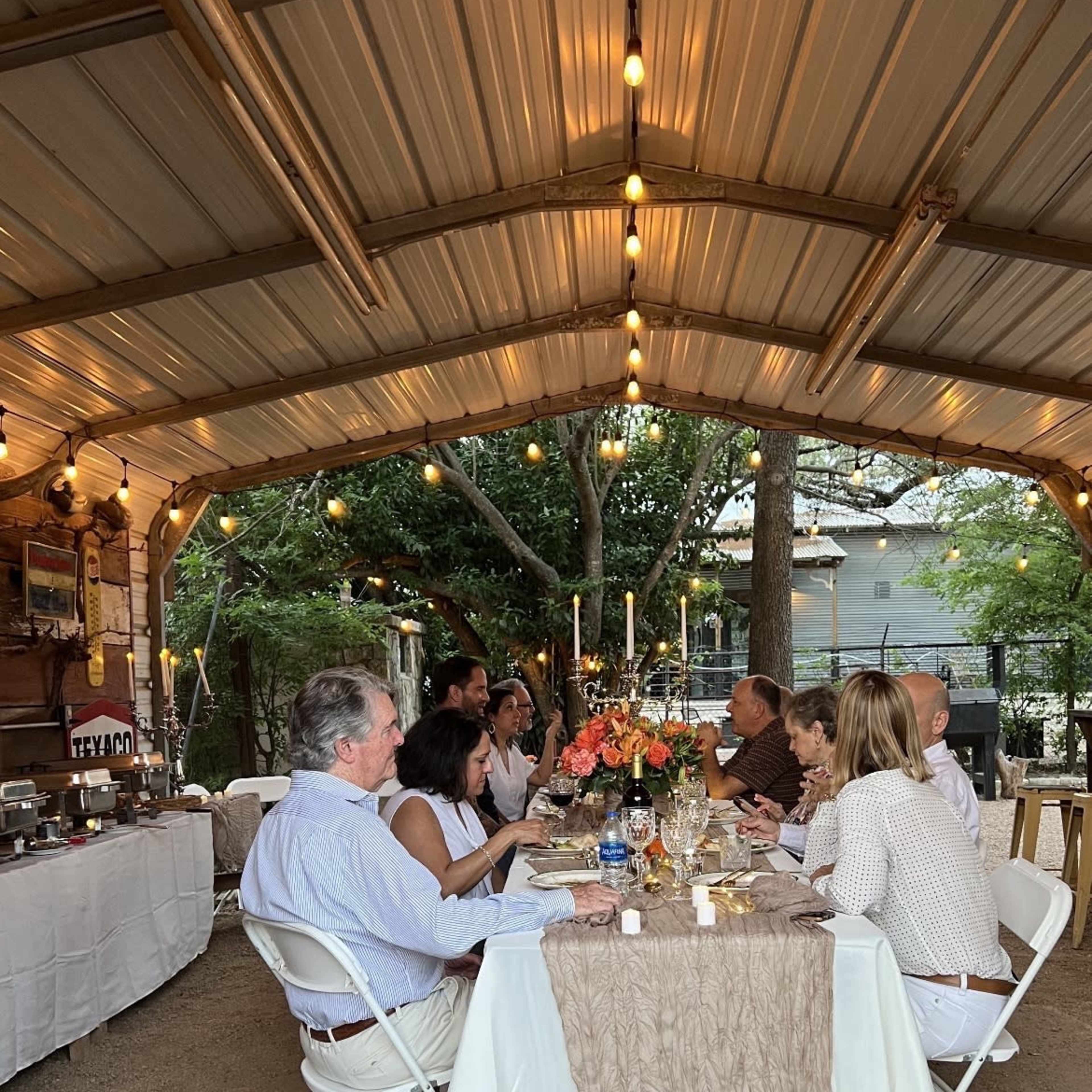 A group of people sits around a table decorated with flowers and candles in a covered outdoor dining area.