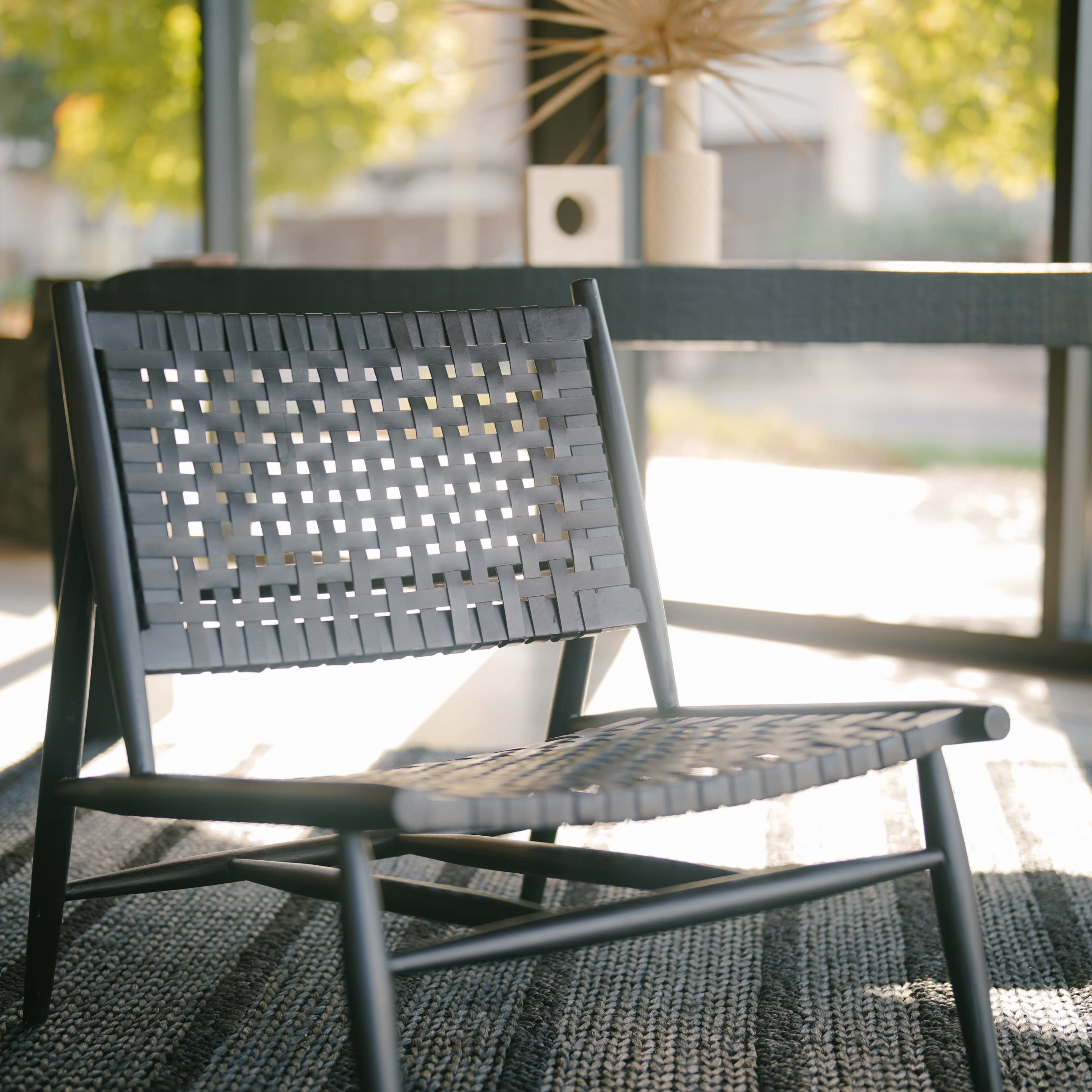 A woven chair is positioned on a textured rug near a table and decorative items, with sunlight streaming through large windows.