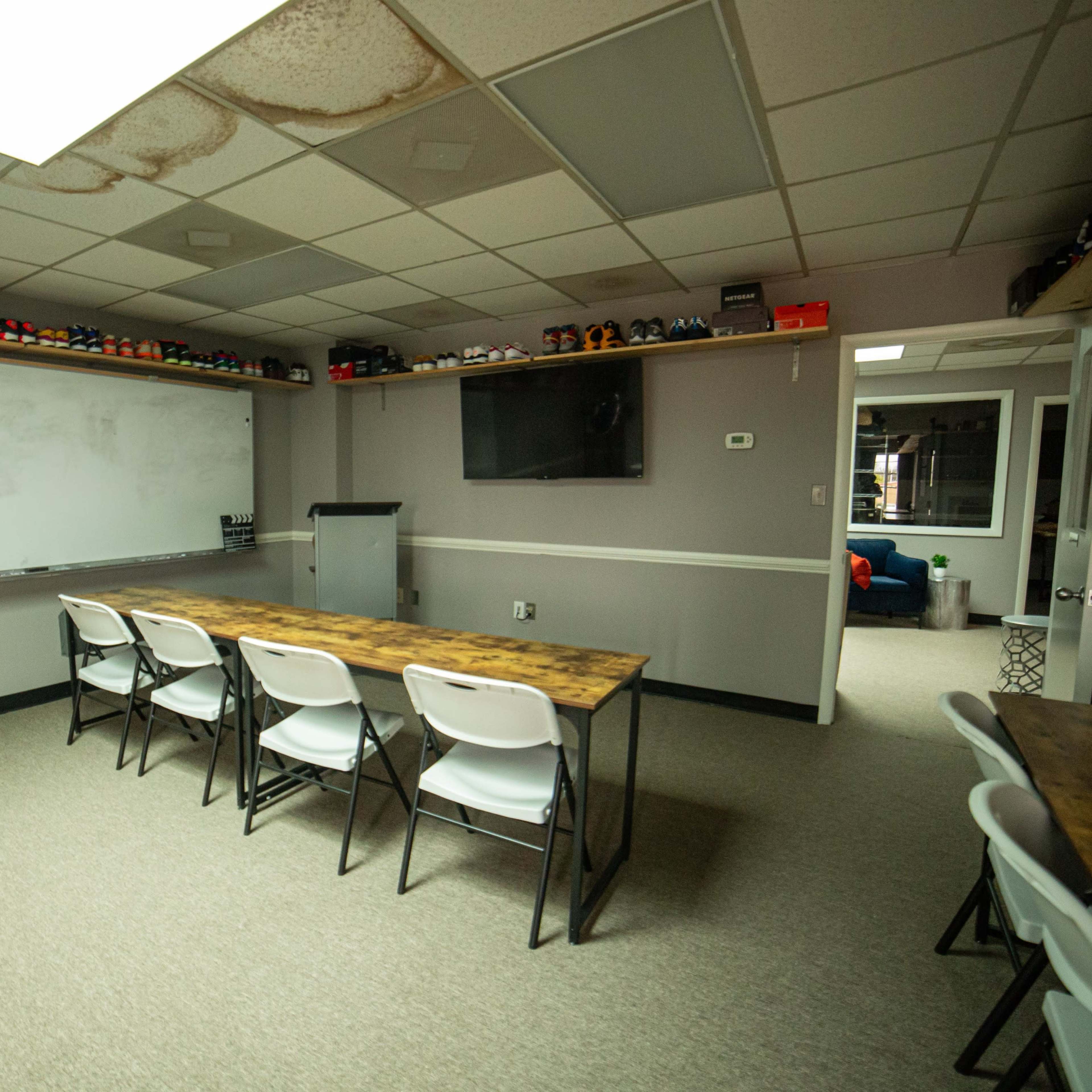 A meeting room with multiple tables and chairs, a whiteboard, a television, and shelves displaying various items.